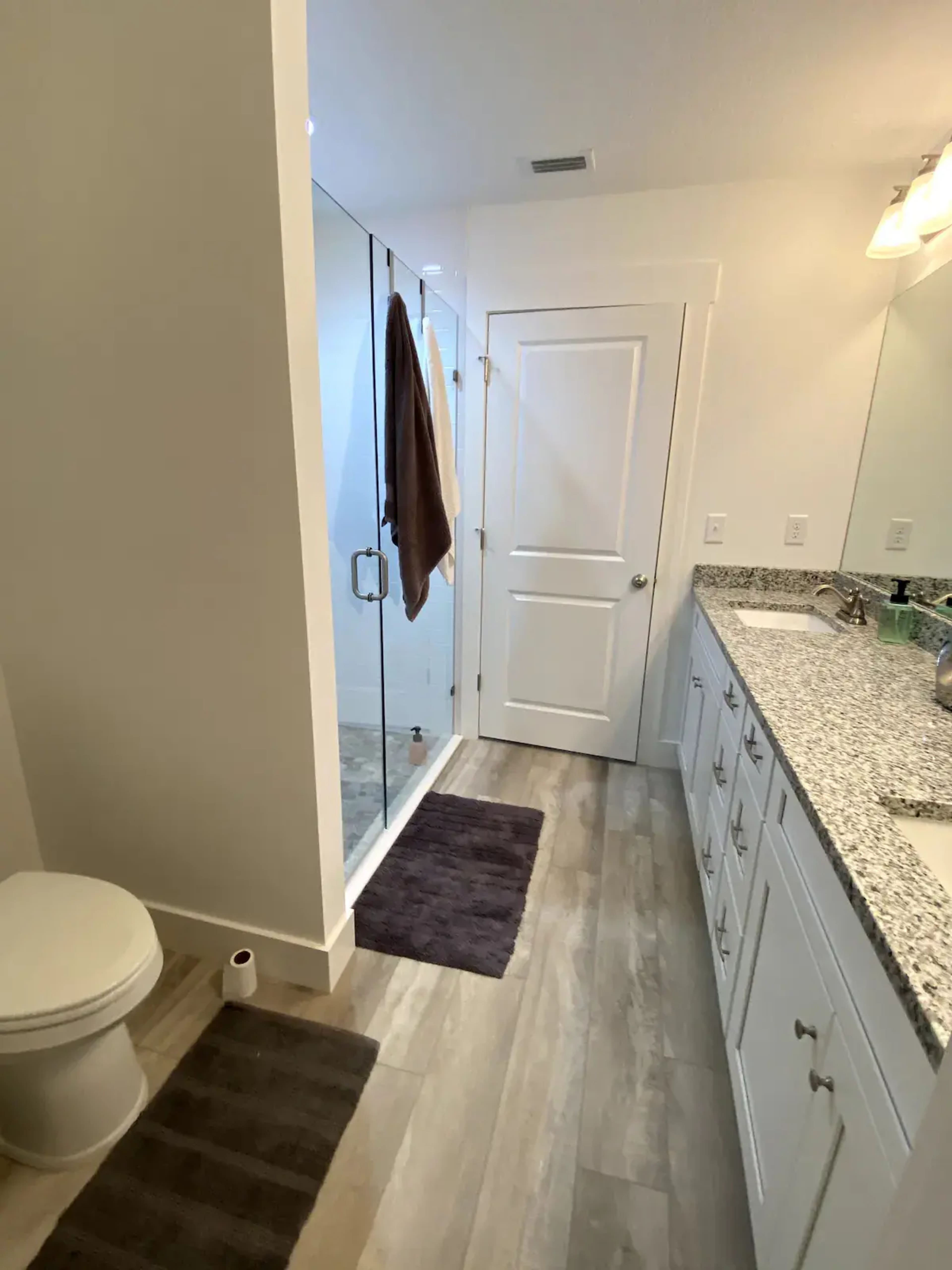 A modern bathroom featuring a glass shower enclosure, a toilet, a double-sink vanity with granite countertops, and light-colored walls and flooring.