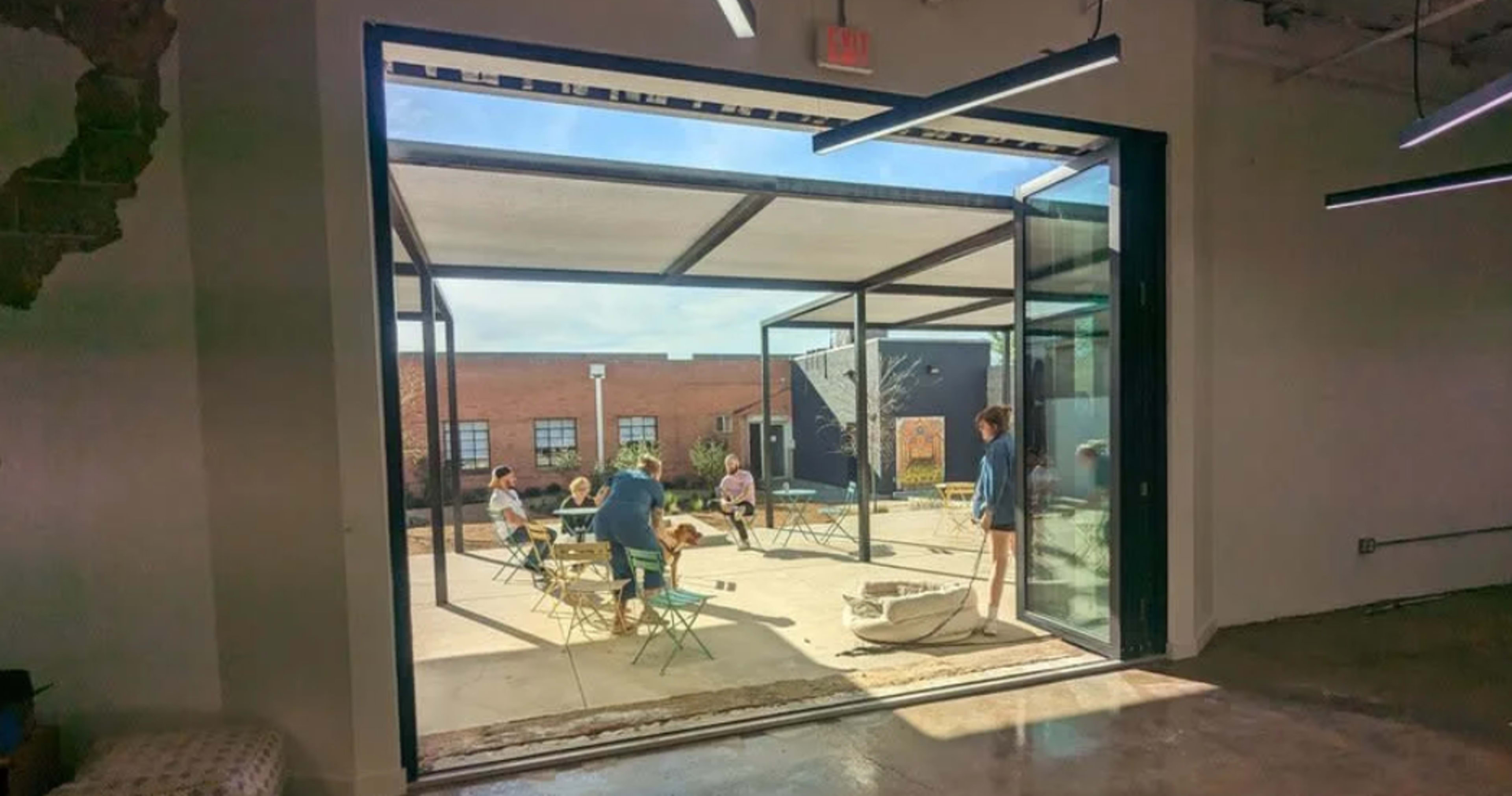 A group of people are sitting at tables in an outdoor space surrounded by buildings, with large glass doors open to the area.