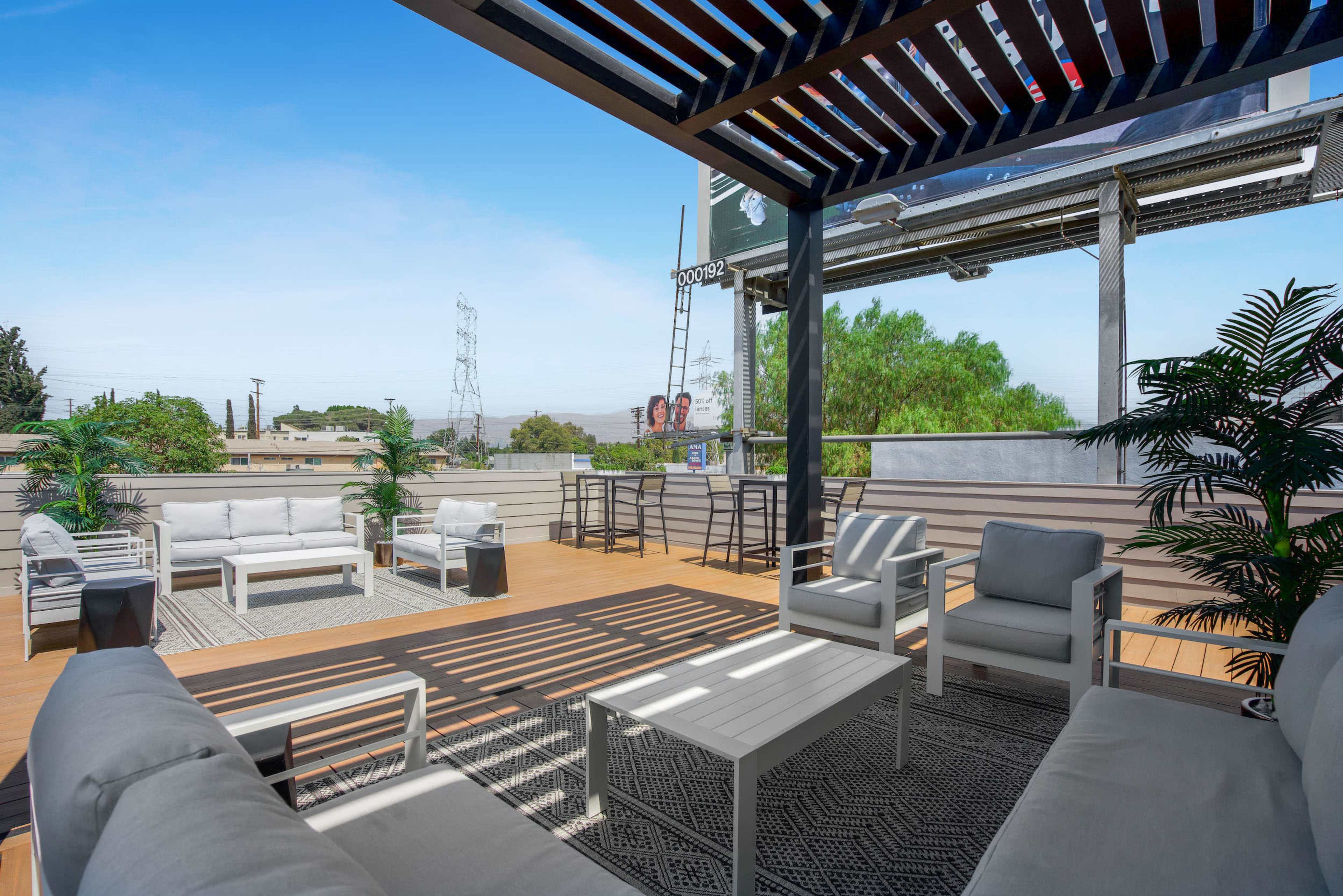 A modern outdoor patio features white furniture, a wooden deck, and a view of greenery and a billboard in the background.