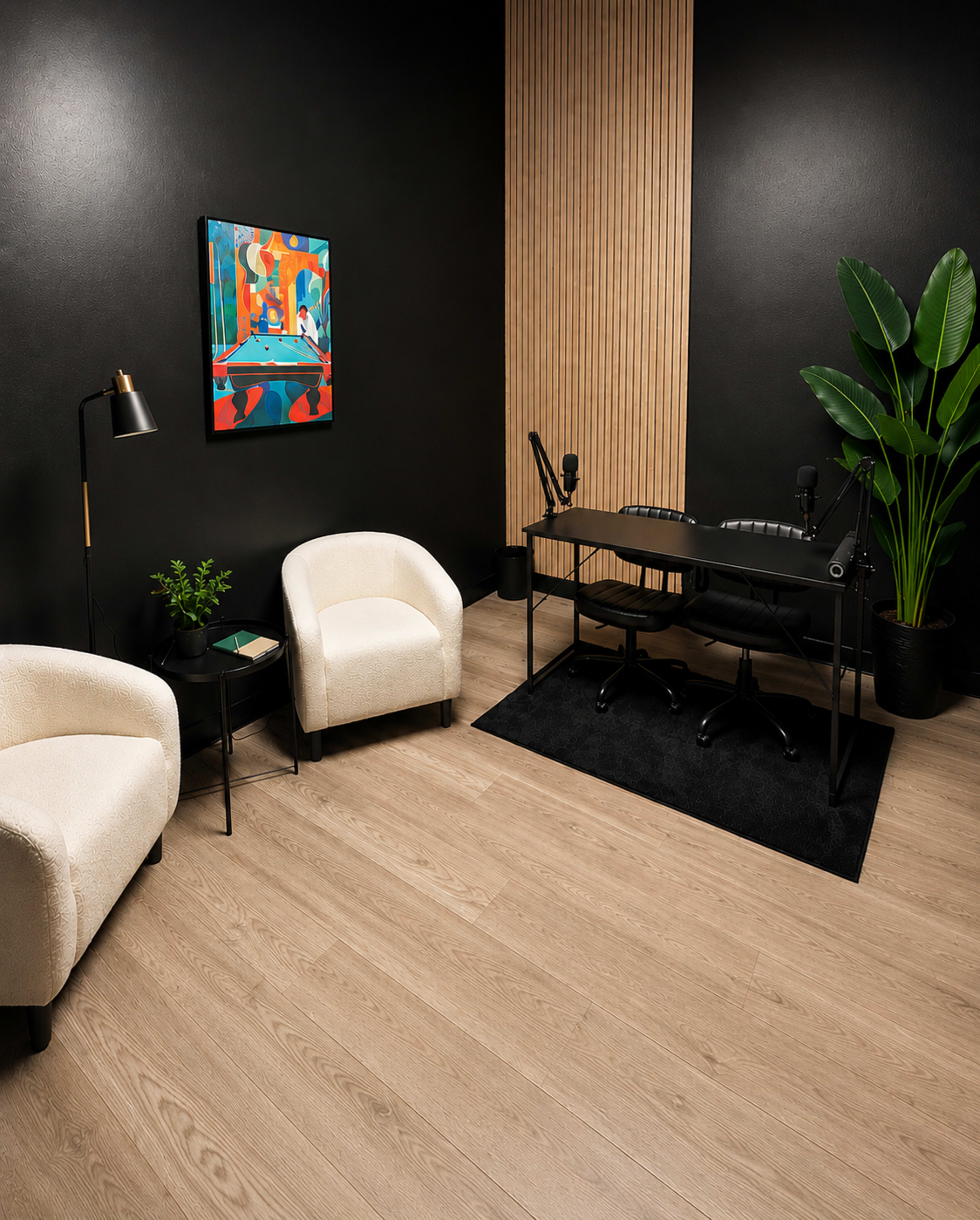 The image shows a modern office space featuring two white lounge chairs, a black desk with microphone stands, a small side table, and a large indoor plant against a black wall.