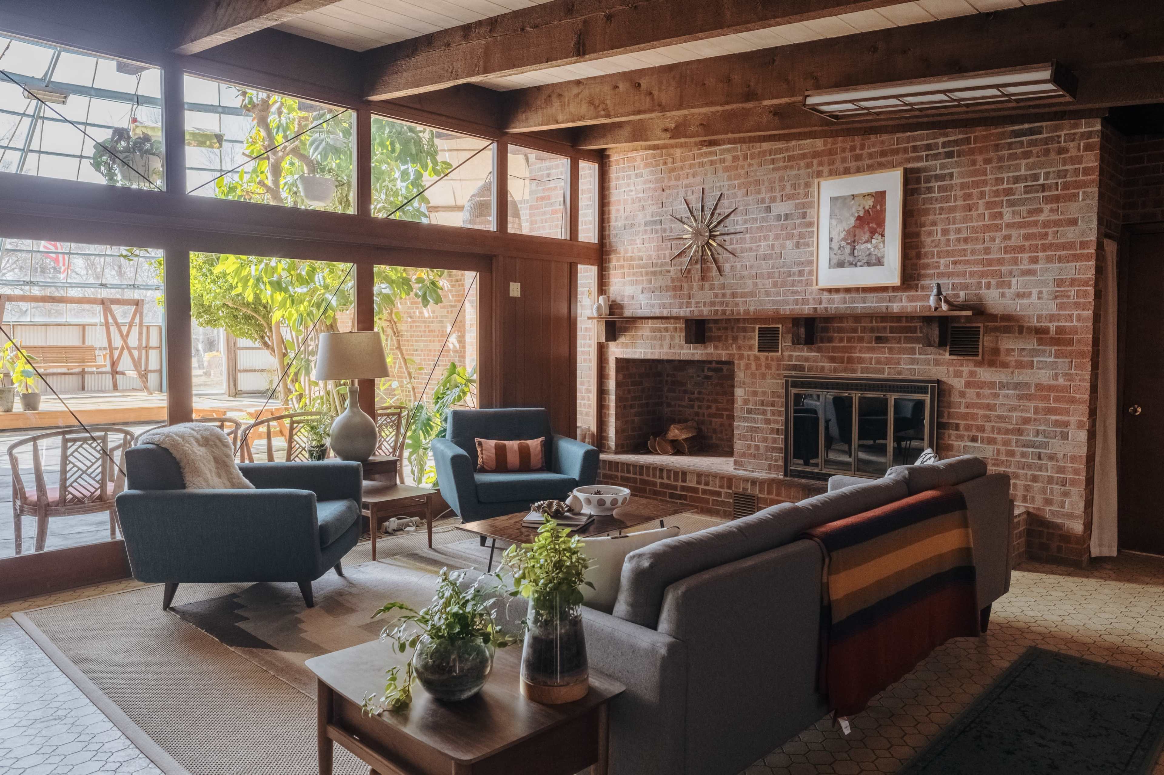 A spacious living room features a brick fireplace, large windows, and a combination of blue and gray furniture, complemented by indoor plants and wooden accents.