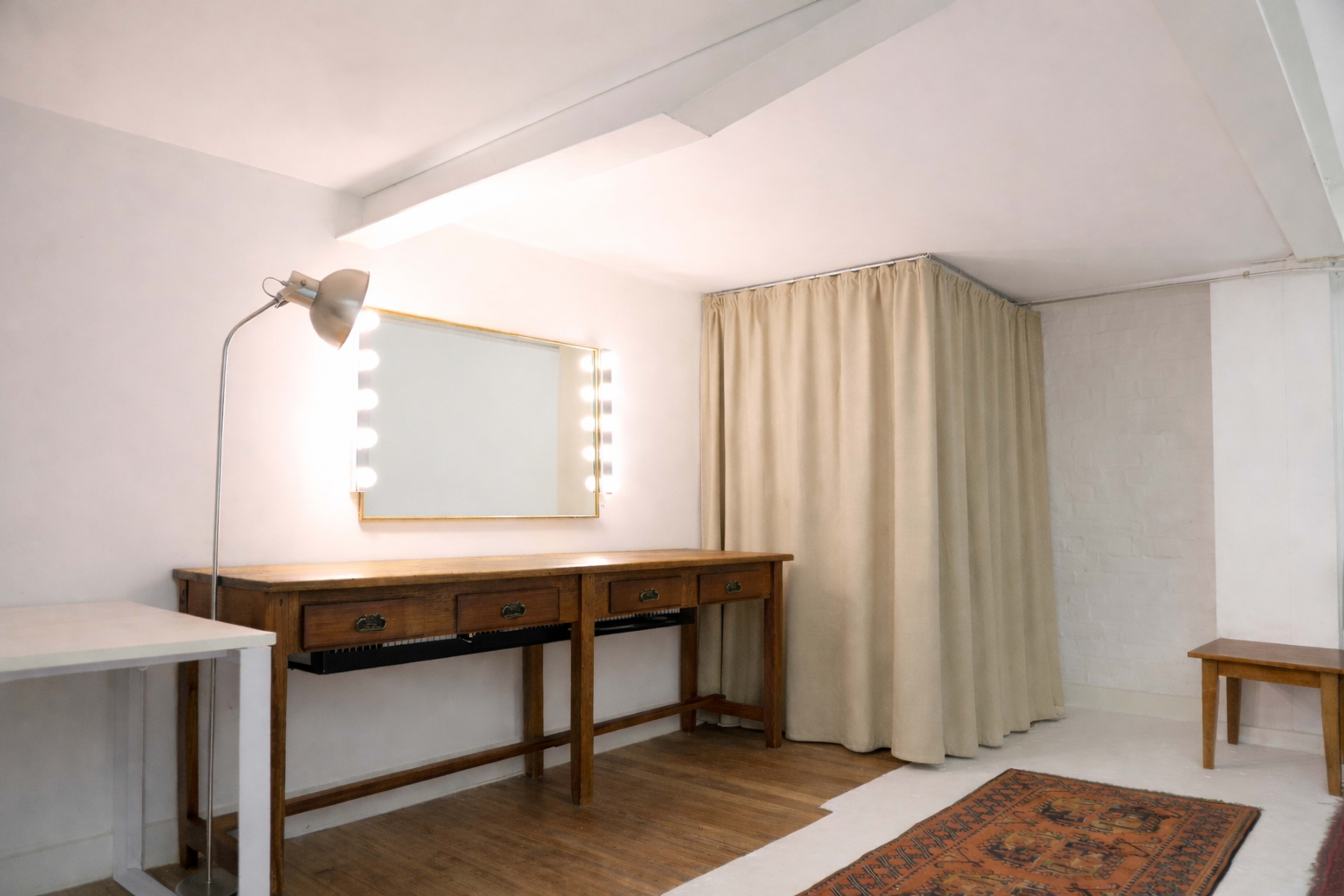 The image shows a simple, well-lit room featuring a wooden vanity table with a mirror, a lamp, a curtain covering a corner, and a patterned rug on the floor.