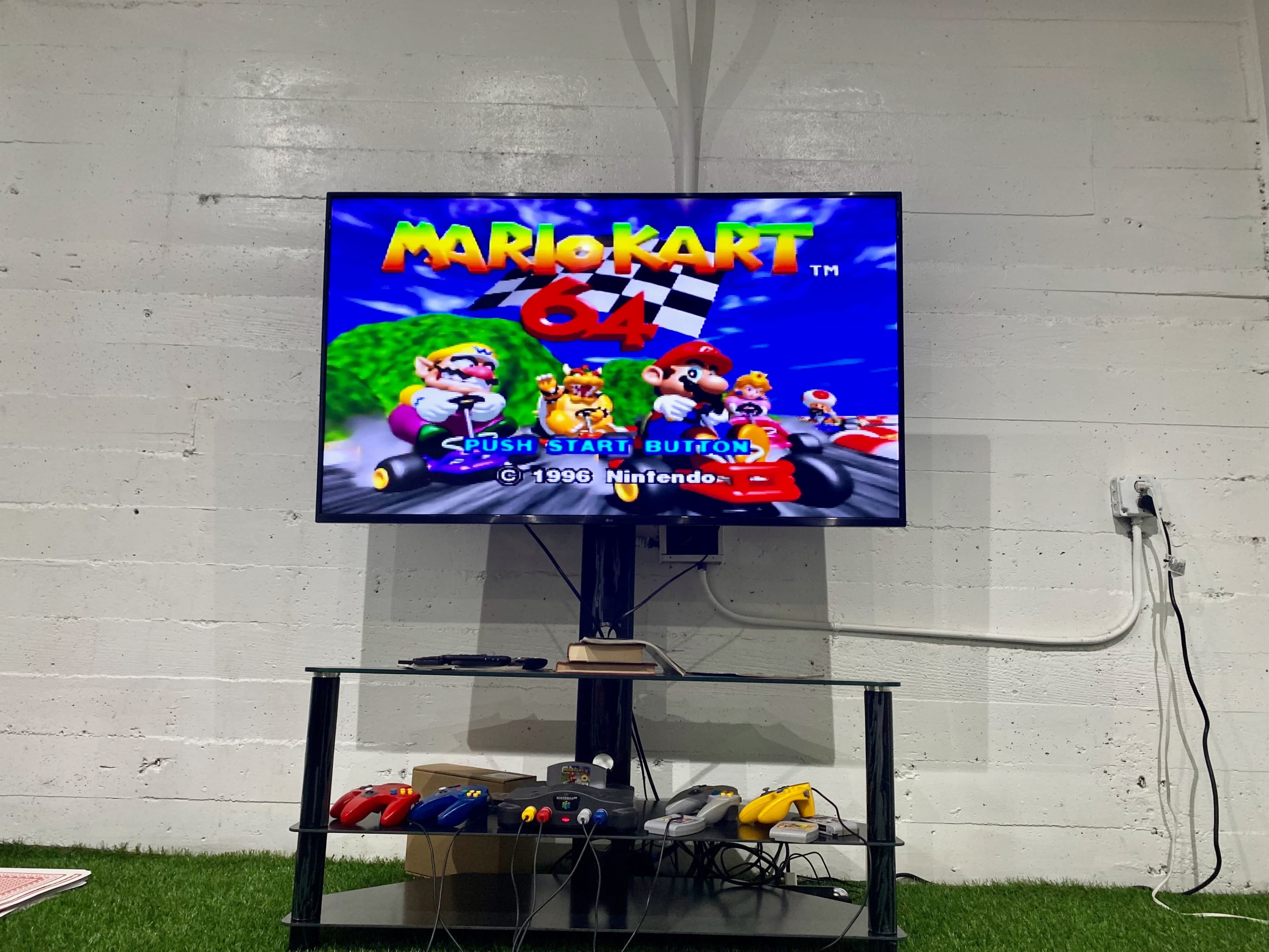 A television displays the Mario Kart 64 title screen on a stand with gaming controllers and accessories beneath it.
