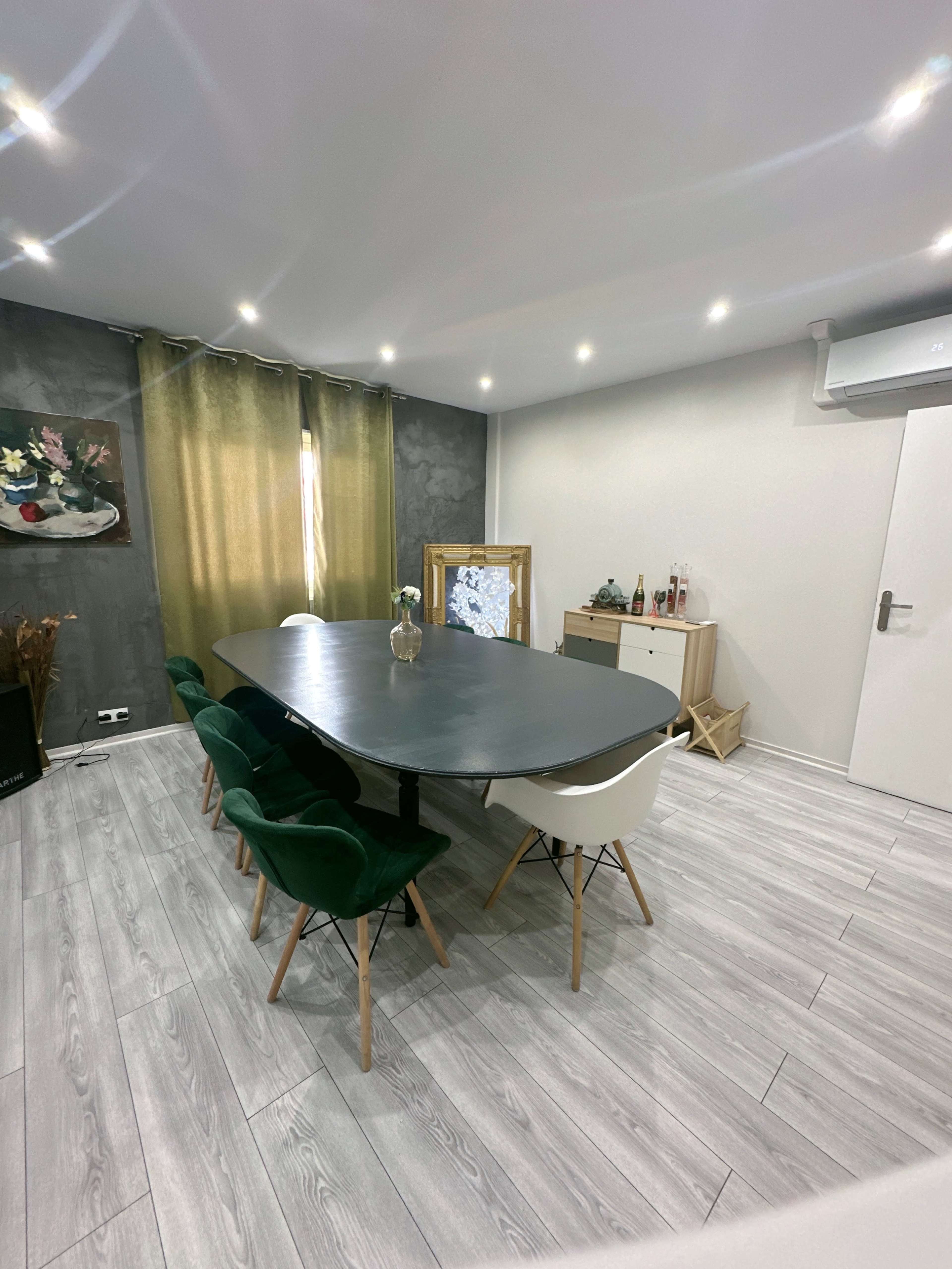 A modern dining area with a large black table surrounded by green and white chairs, complemented by green curtains and minimalist decor.