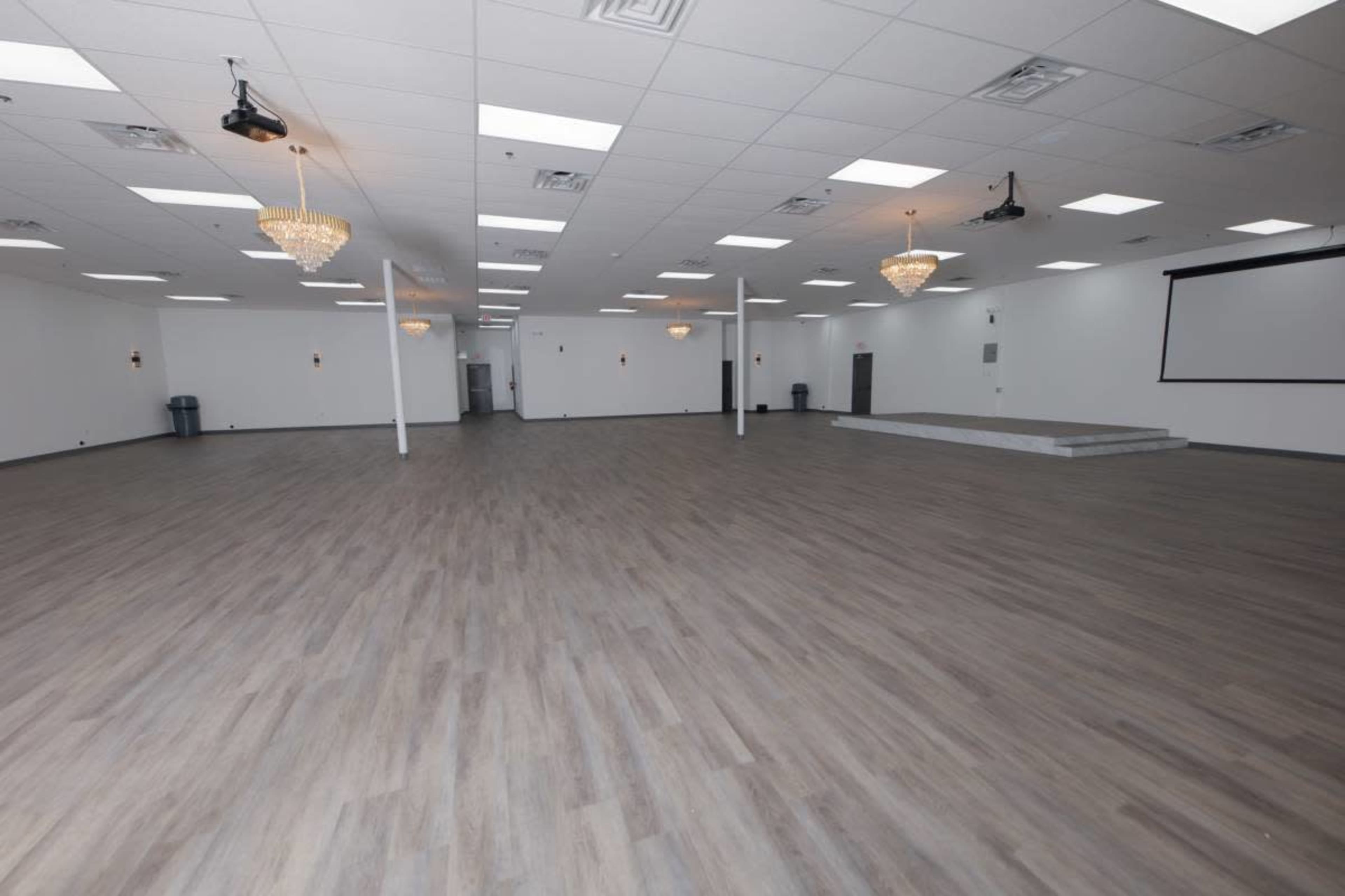 The image shows a large, empty event space with wooden flooring, white walls, and several chandeliers hanging from the ceiling.