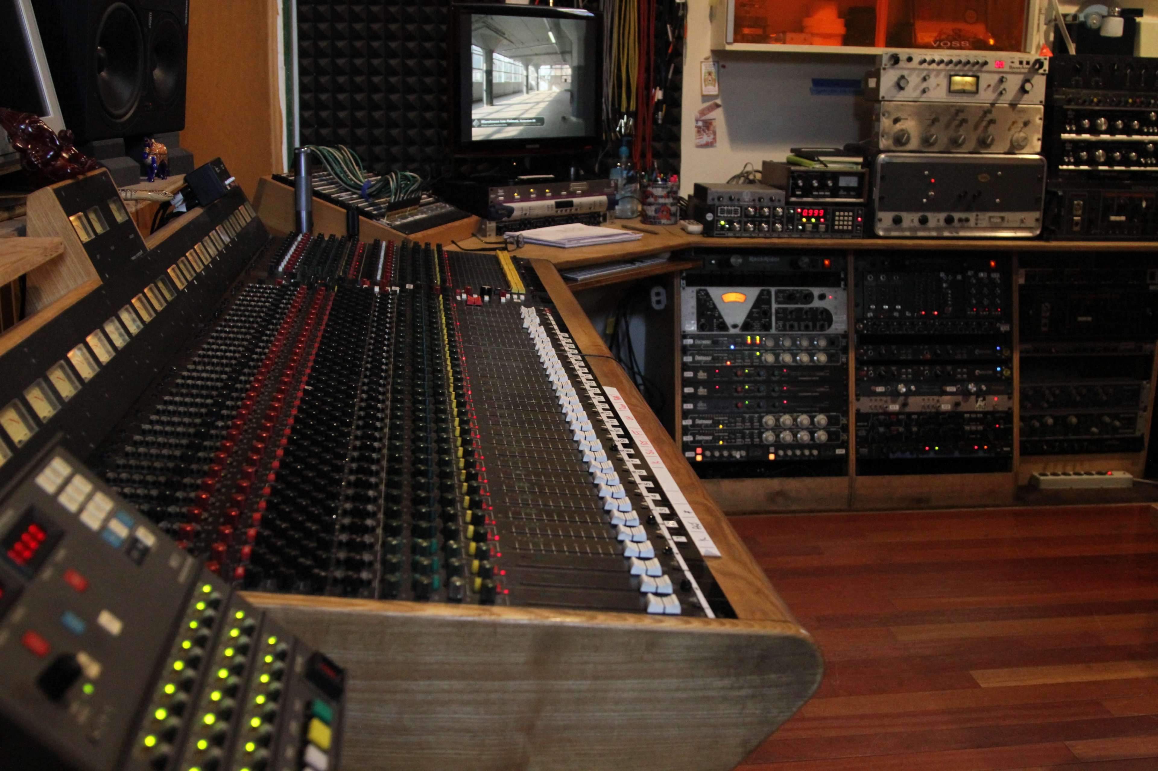 The image shows a recording studio with a large mixing console, various audio equipment, and a television monitor displayed in the background.