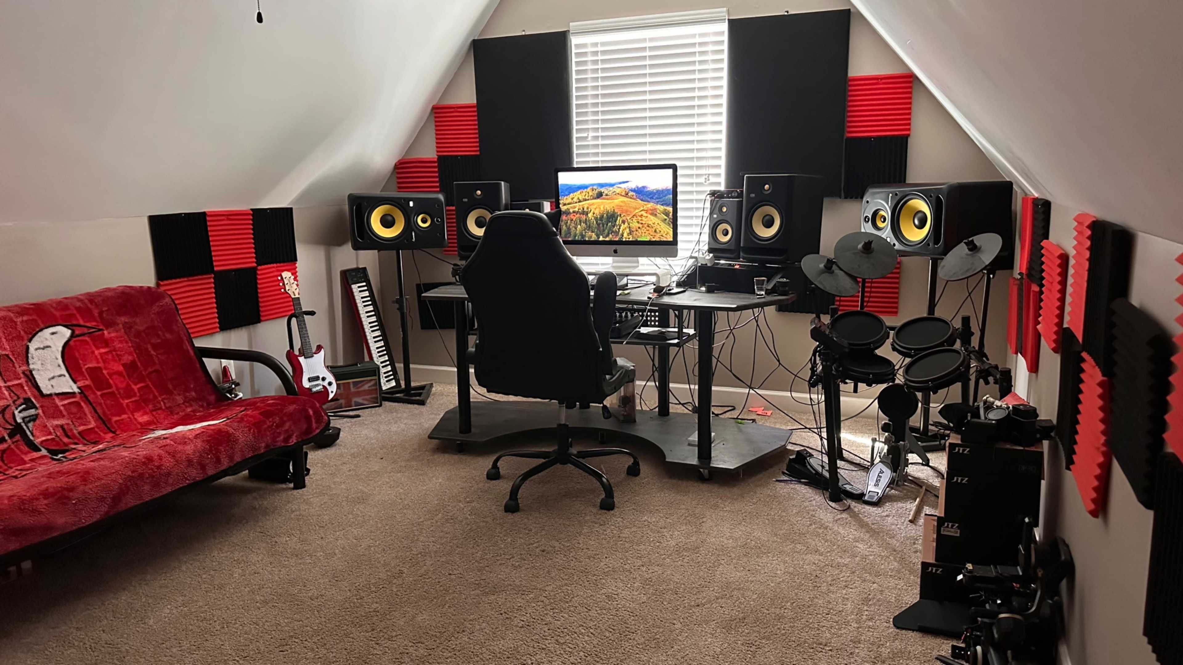 The image shows a home music studio with a desk, computer, monitors, keyboard, electric guitar, and electronic drum set arranged in a room featuring red and black acoustic panels.