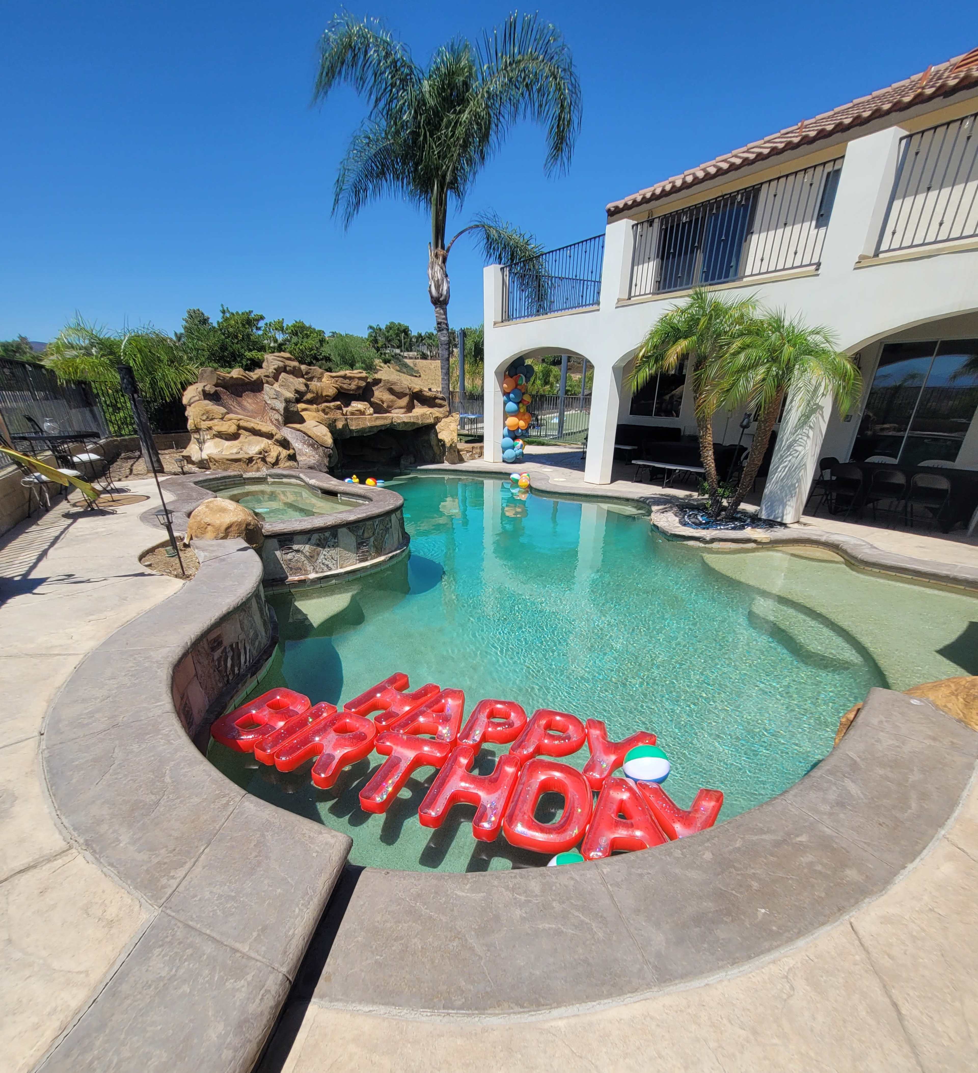 A swimming pool with "HAPPY BIRTHDAY" balloons floats on the water, surrounded by palm trees and a stone waterfall.