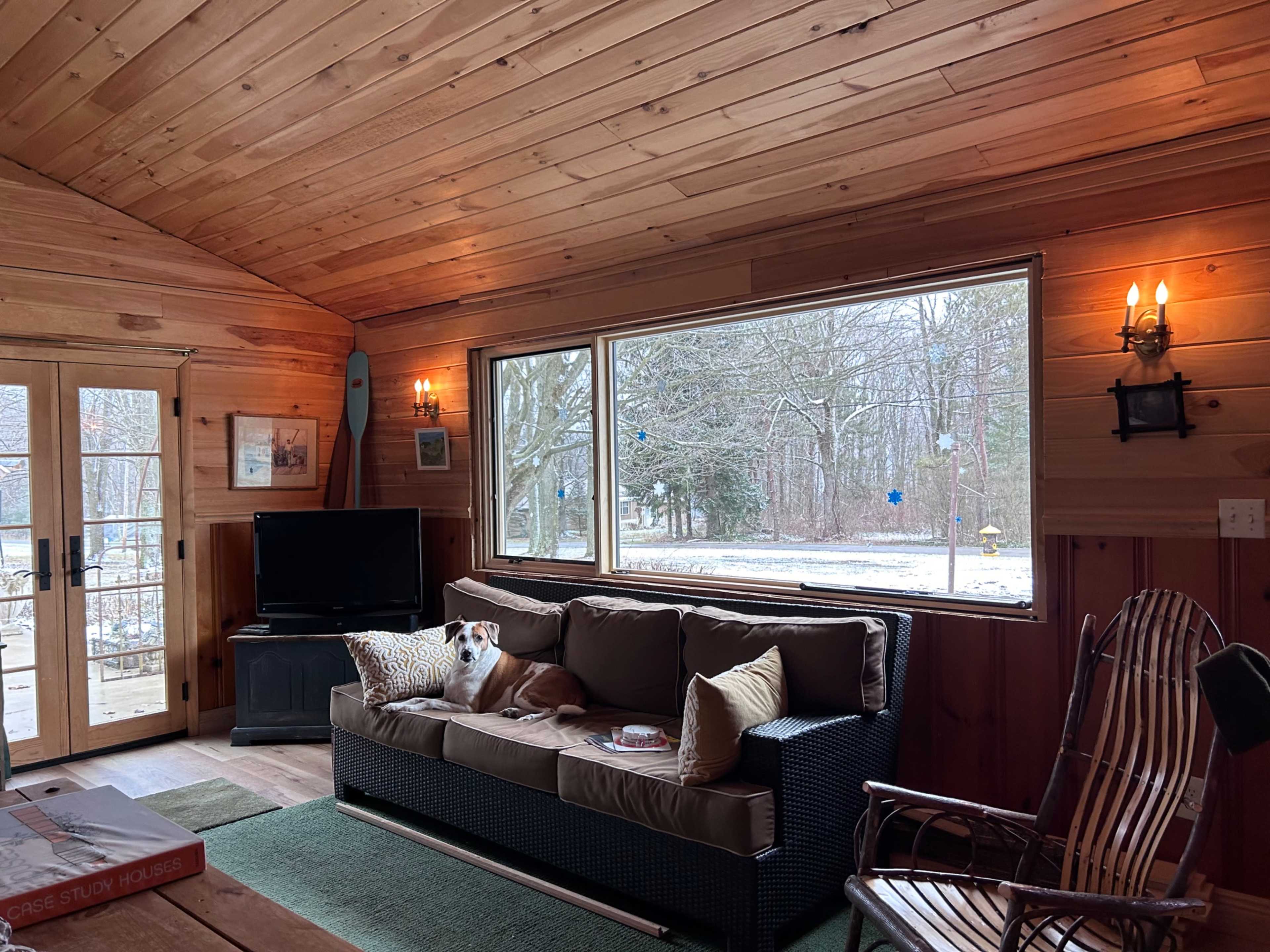 A cozy living room features a wooden ceiling, a sectional couch, a TV, and large windows overlooking a snowy outdoor scene.