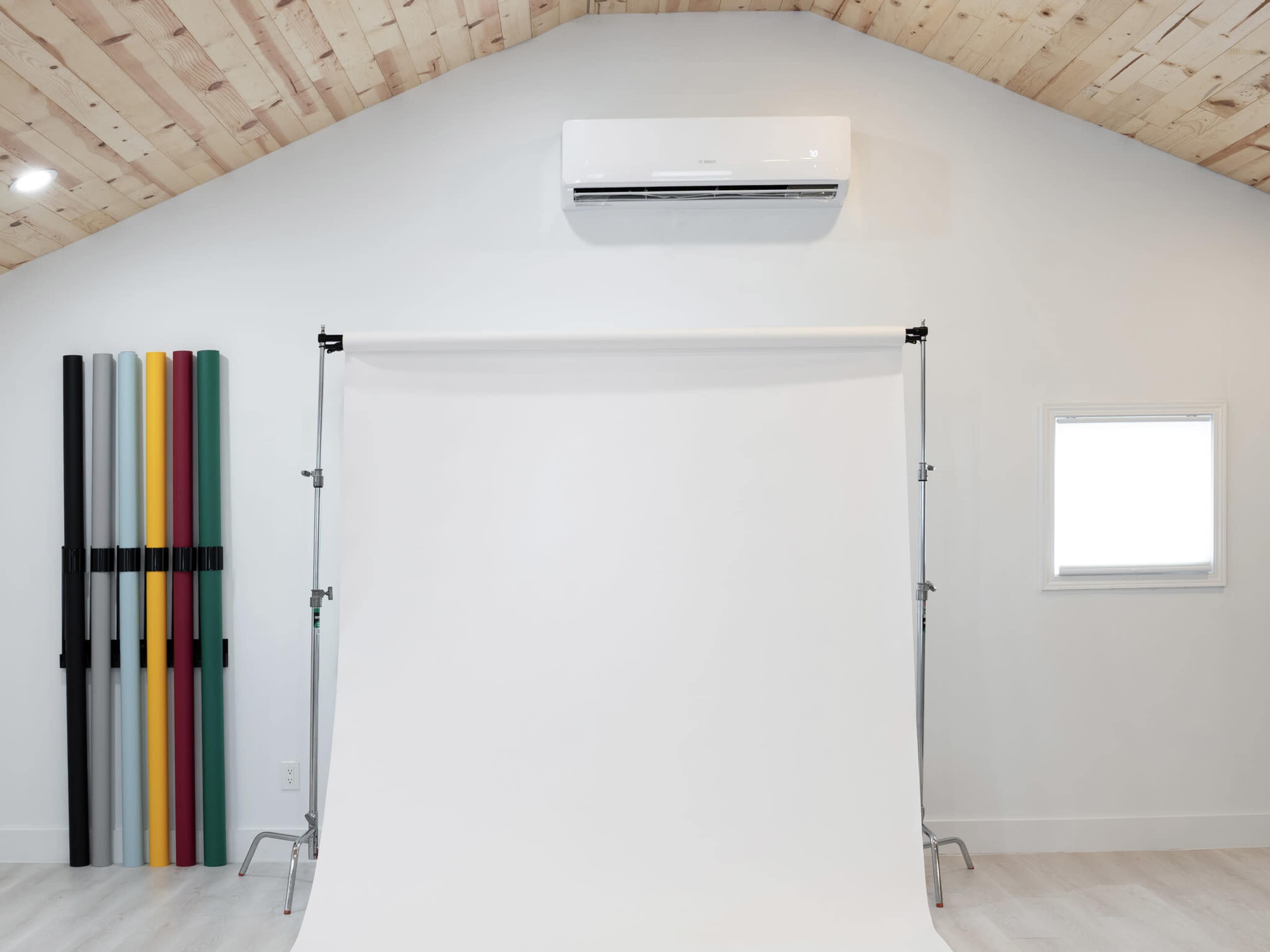 A photography studio with a white backdrop, wooden ceiling, and colored backdrop rolls arranged on the side.
