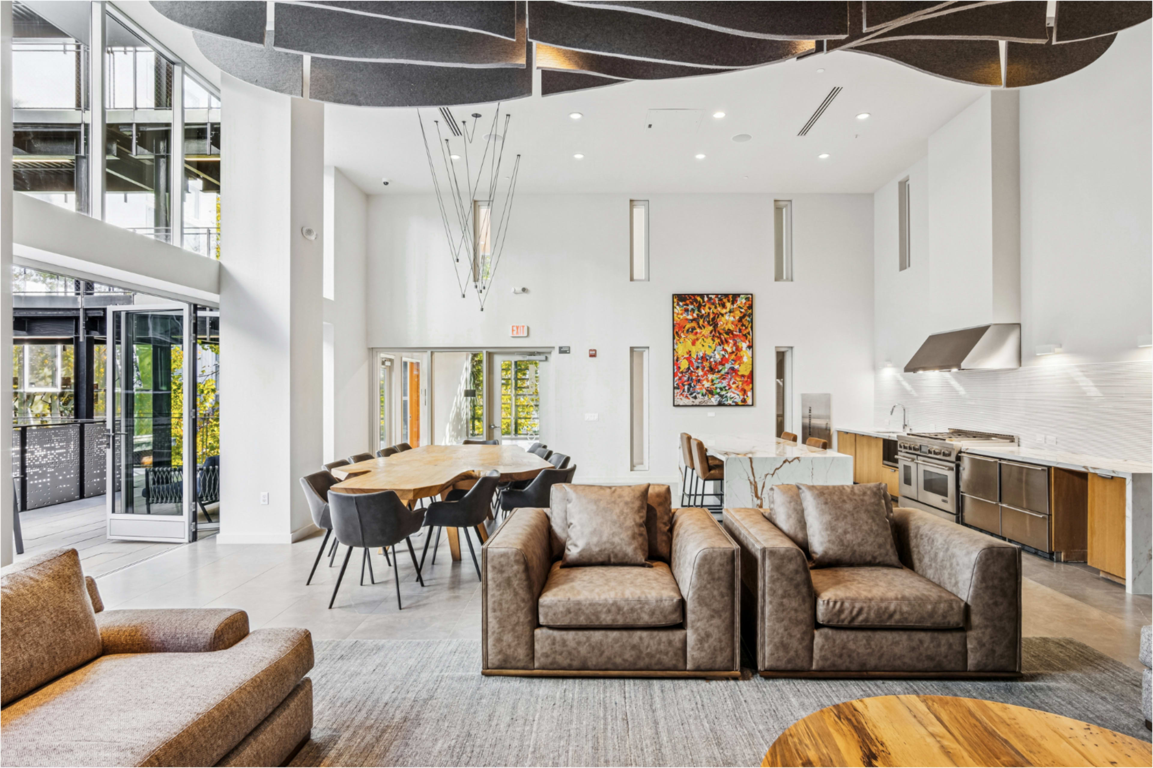 The image shows a modern, open-concept communal area featuring two brown sofas, a large dining table with black chairs, and a kitchen area, all illuminated by natural light through large windows.