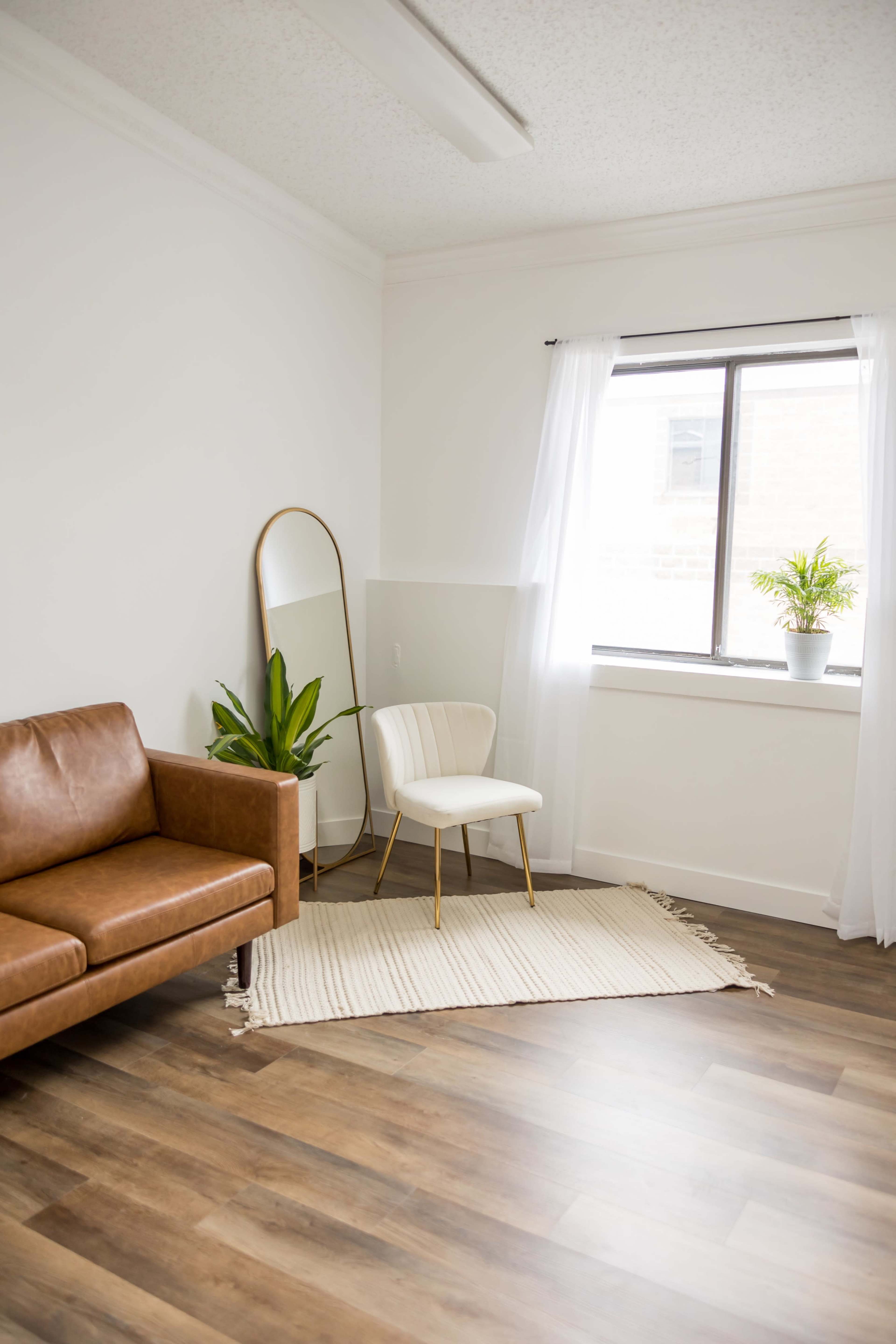 A minimalist living space featuring a brown leather sofa, a light-colored chair, a large mirror, a potted plant, and a window with sheer curtains.