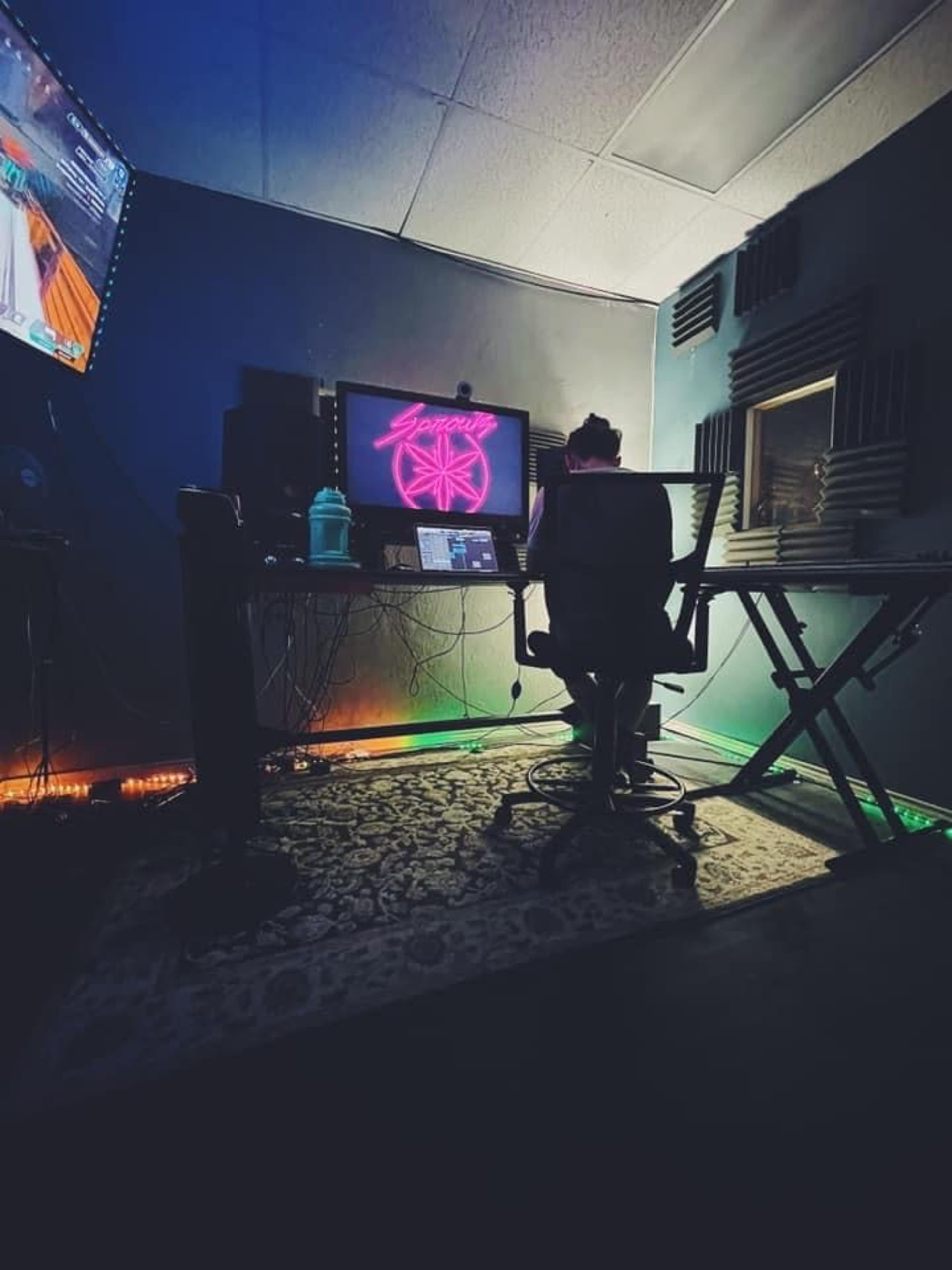 A person sits at a desk with a computer and colorful lighting in a dimly lit room that features soundproofing panels.