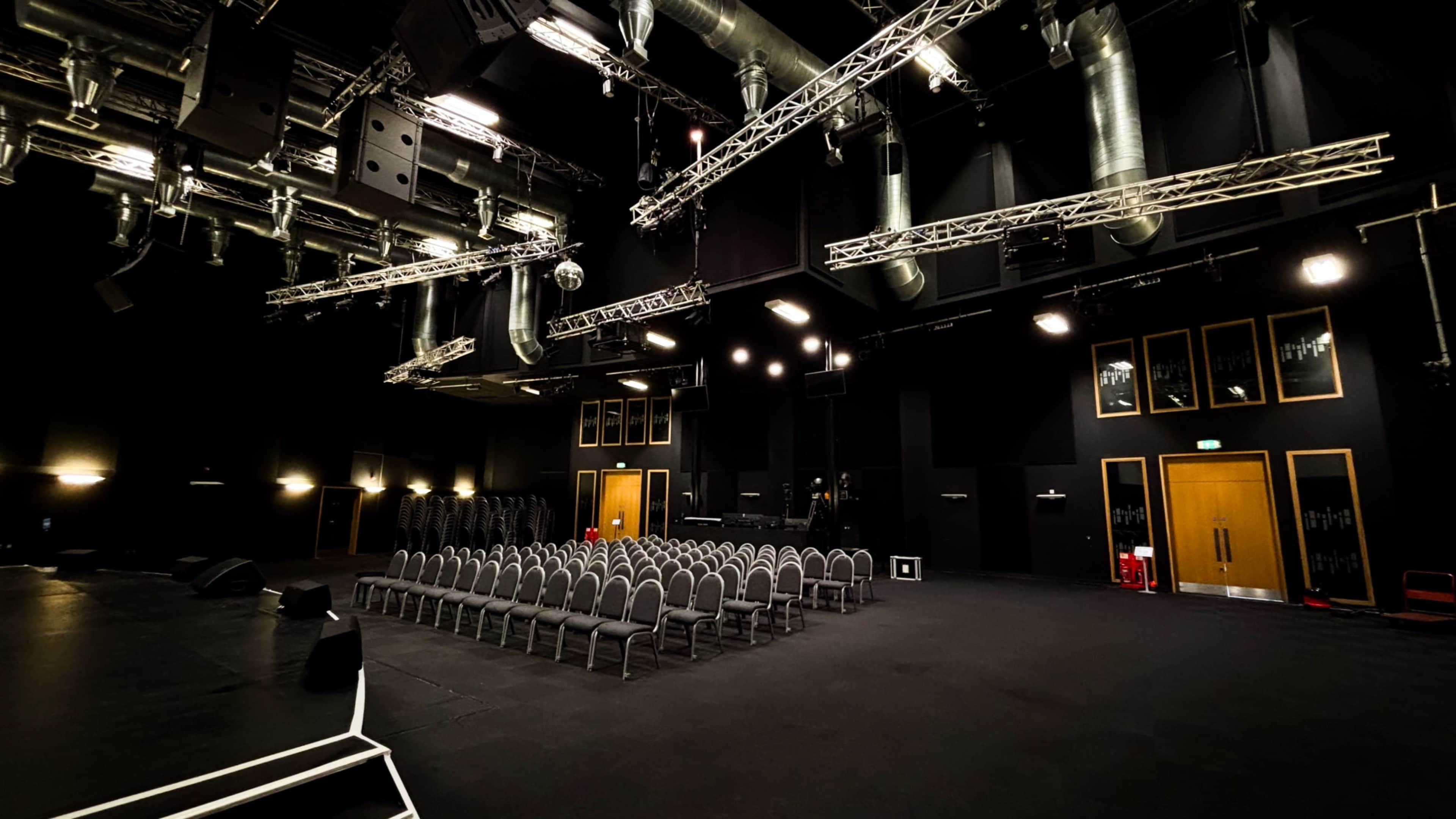 The image shows a dark, spacious auditorium with neatly arranged chairs, sound equipment, and overhead lighting.