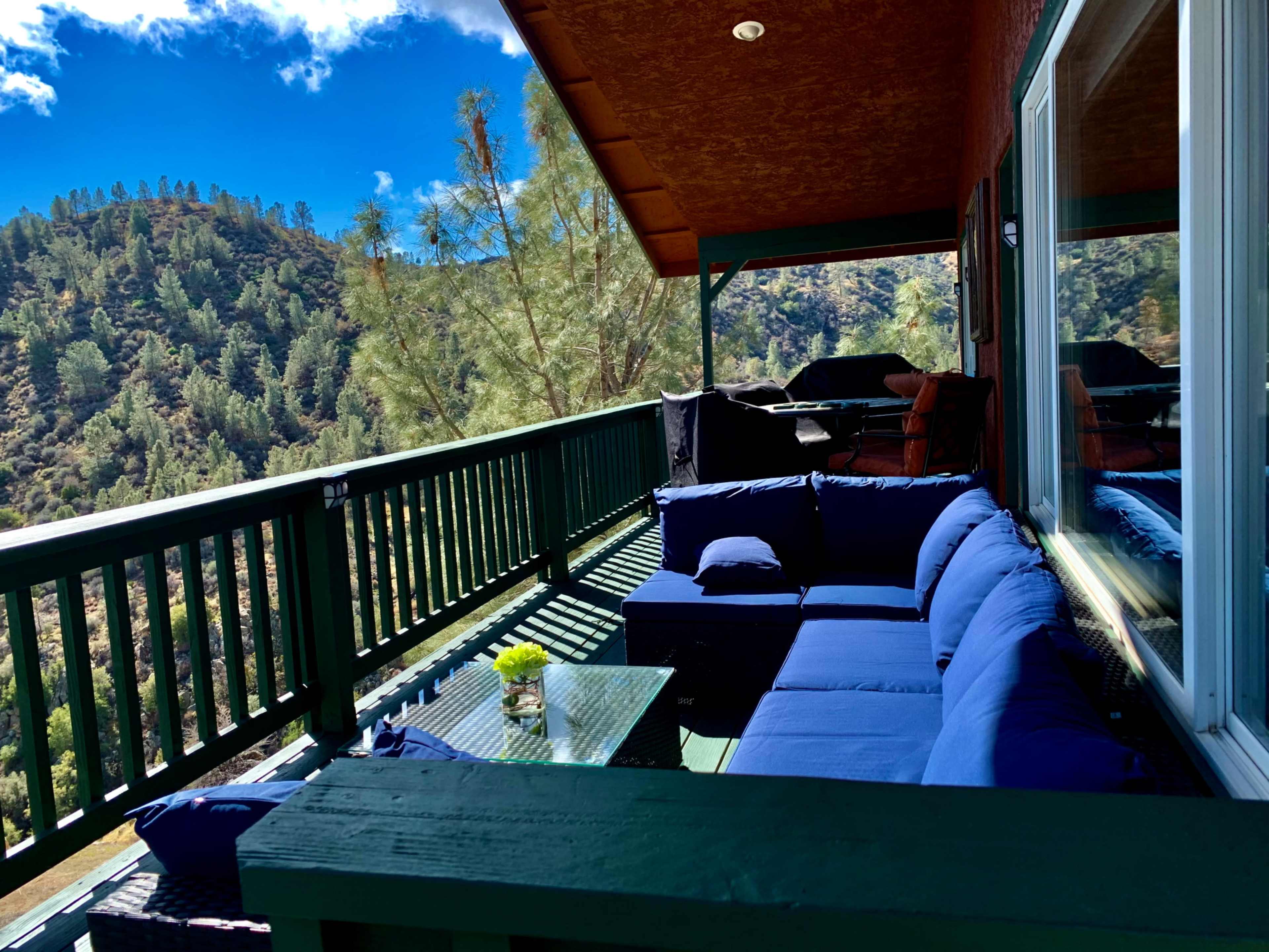 A balcony features a seating area with blue cushions and a small table, overlooking a mountainous landscape under a clear blue sky.