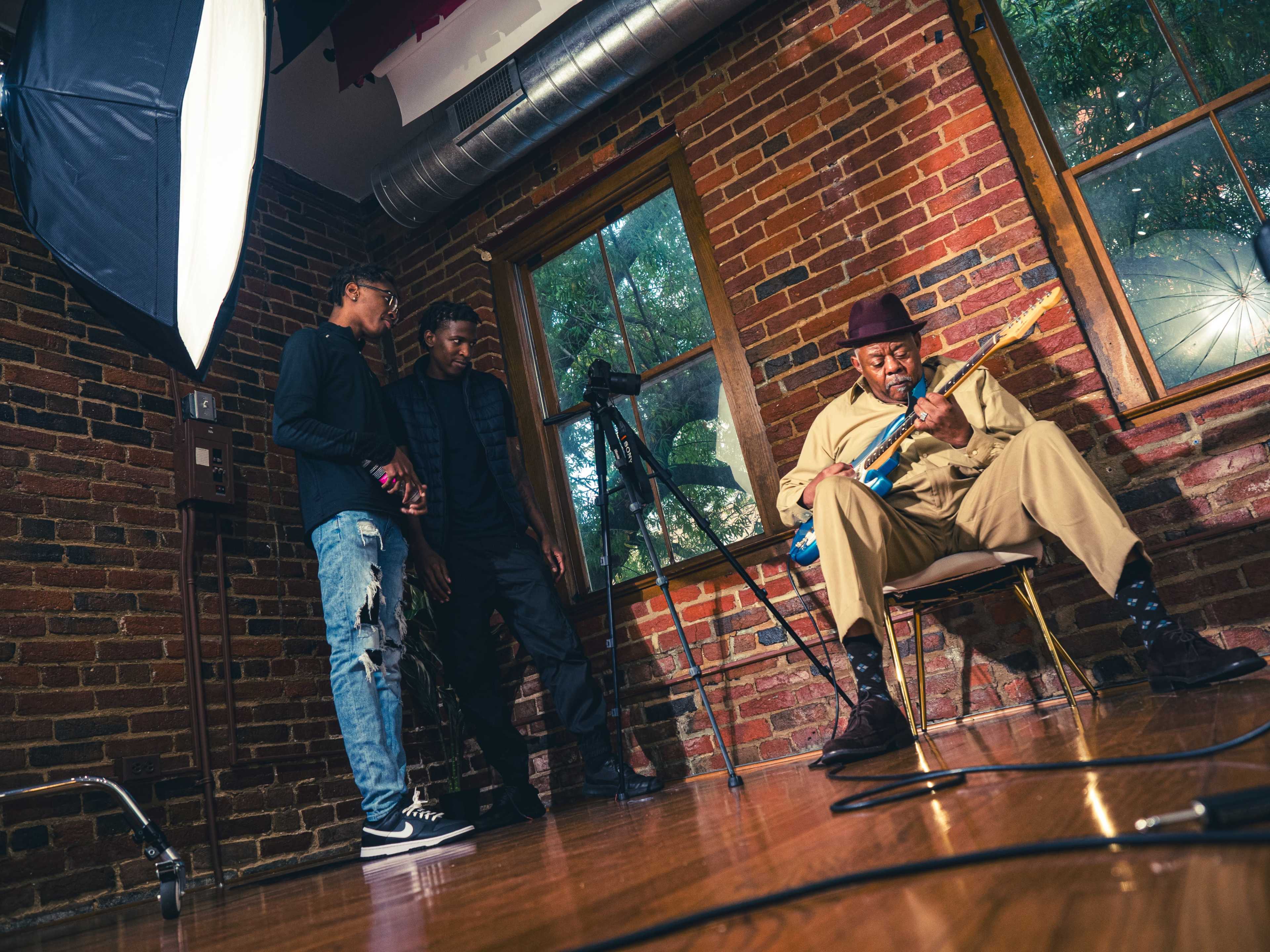 A musician plays an electric guitar while seated in a chair, with two spectators standing nearby in a brick-walled room.