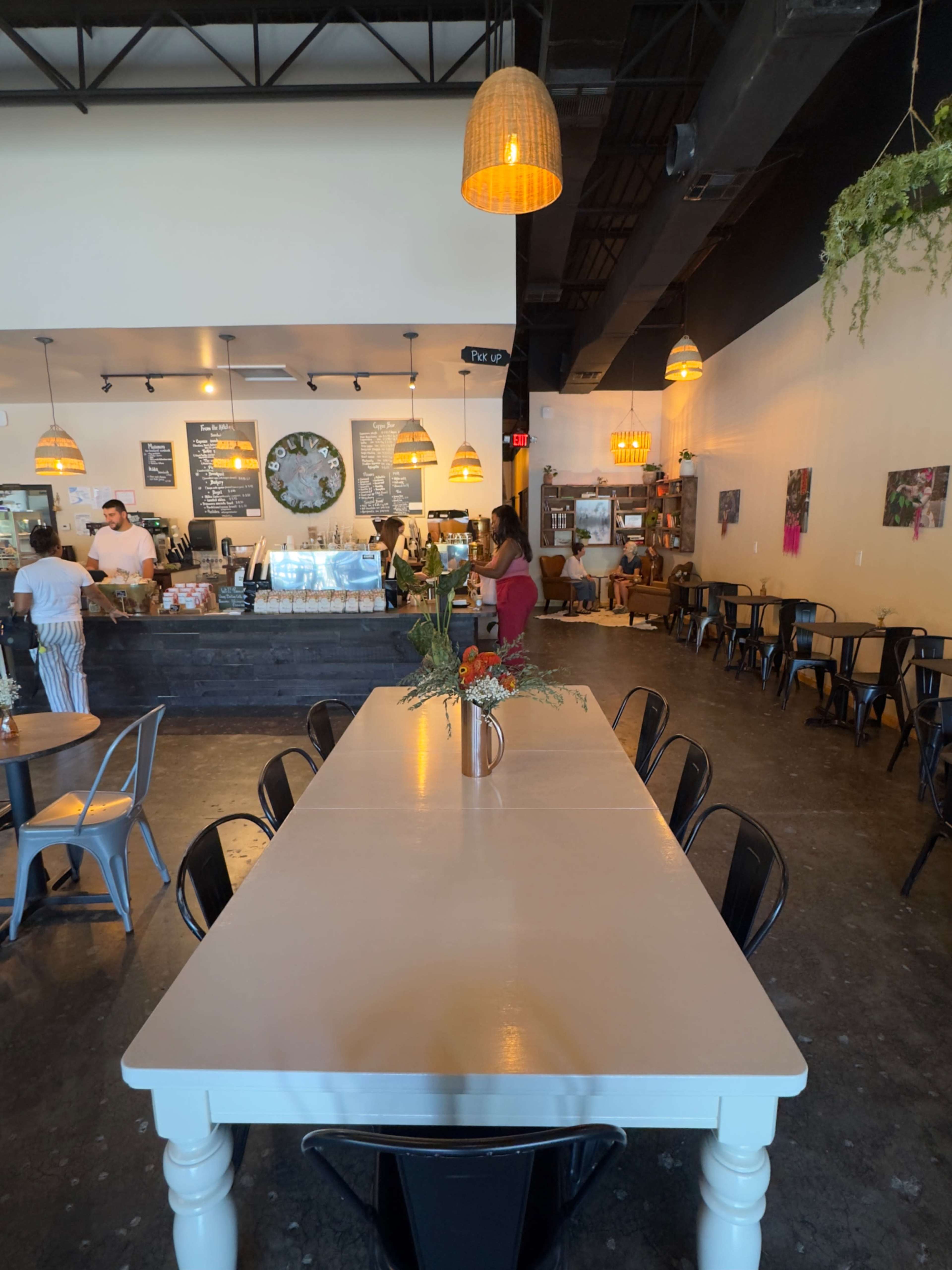 The image shows the interior of a café featuring a large communal table in the foreground, with a counter displaying food and drinks in the background.