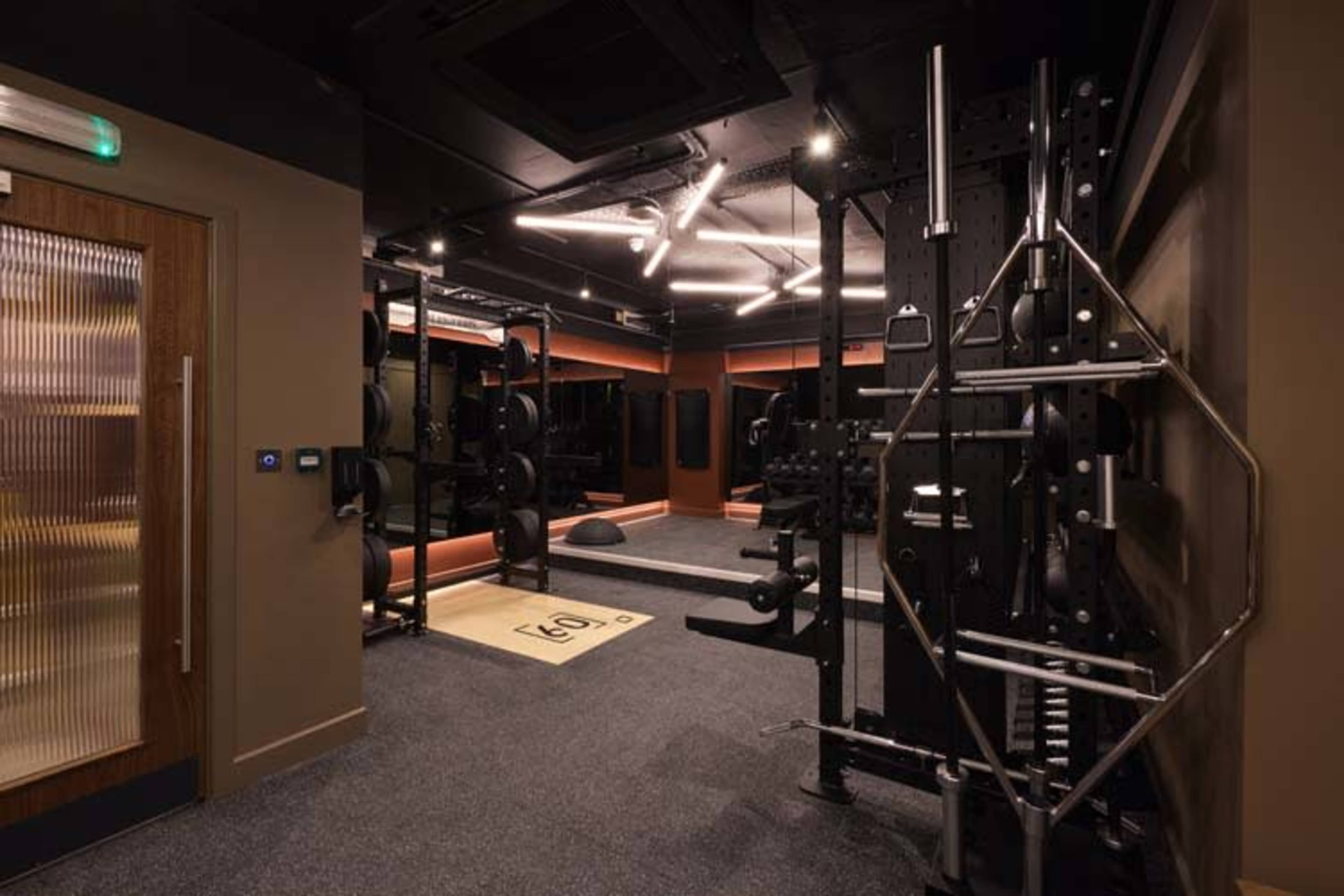 The image shows a modern gym interior featuring a weightlifting area with various equipment, including a squat rack and weight plates, surrounded by dark walls and flooring.