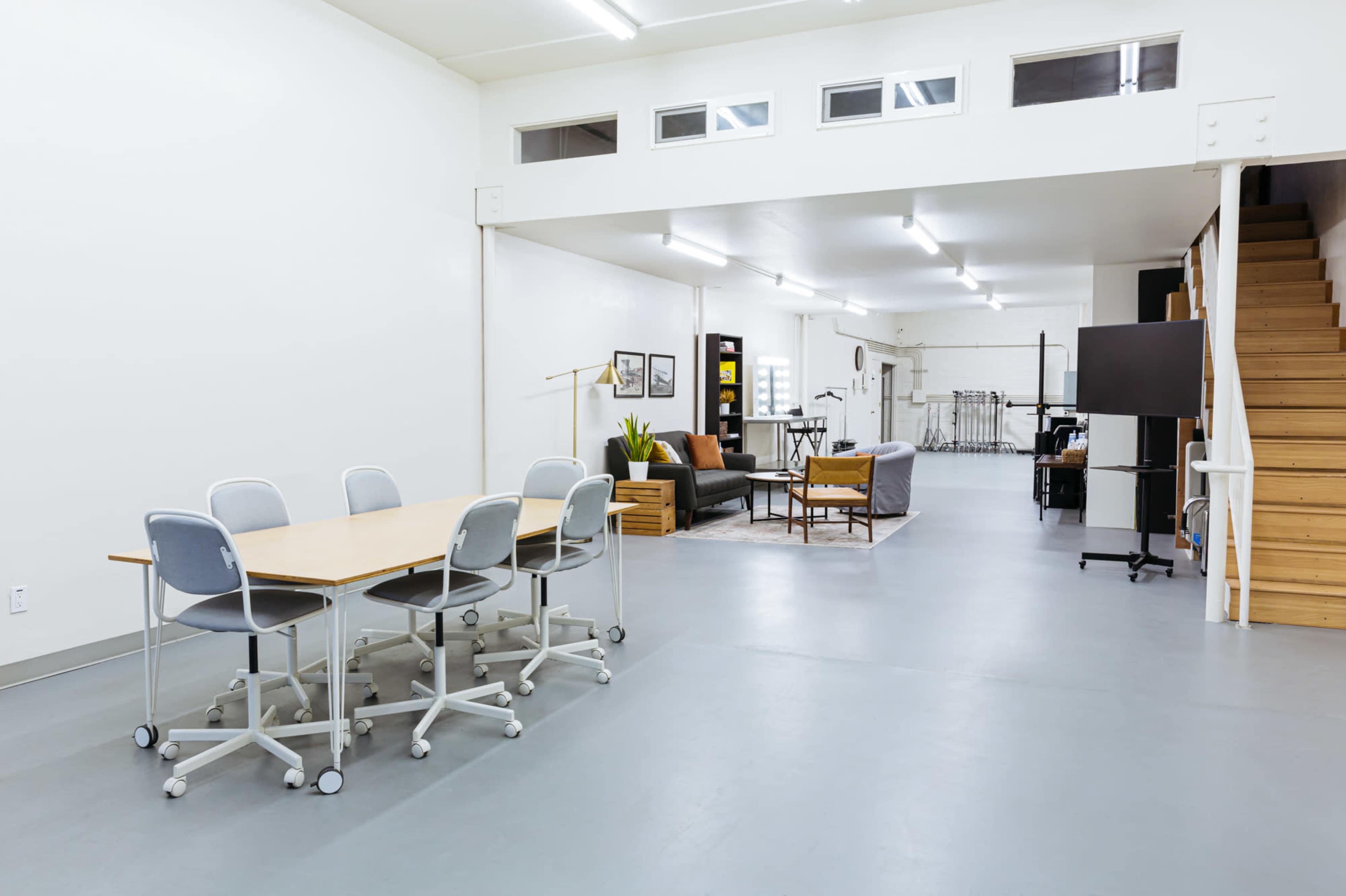 A spacious, well-lit interior featuring a meeting area with a table and chairs on one side and a living space with seating and a staircase on the other.