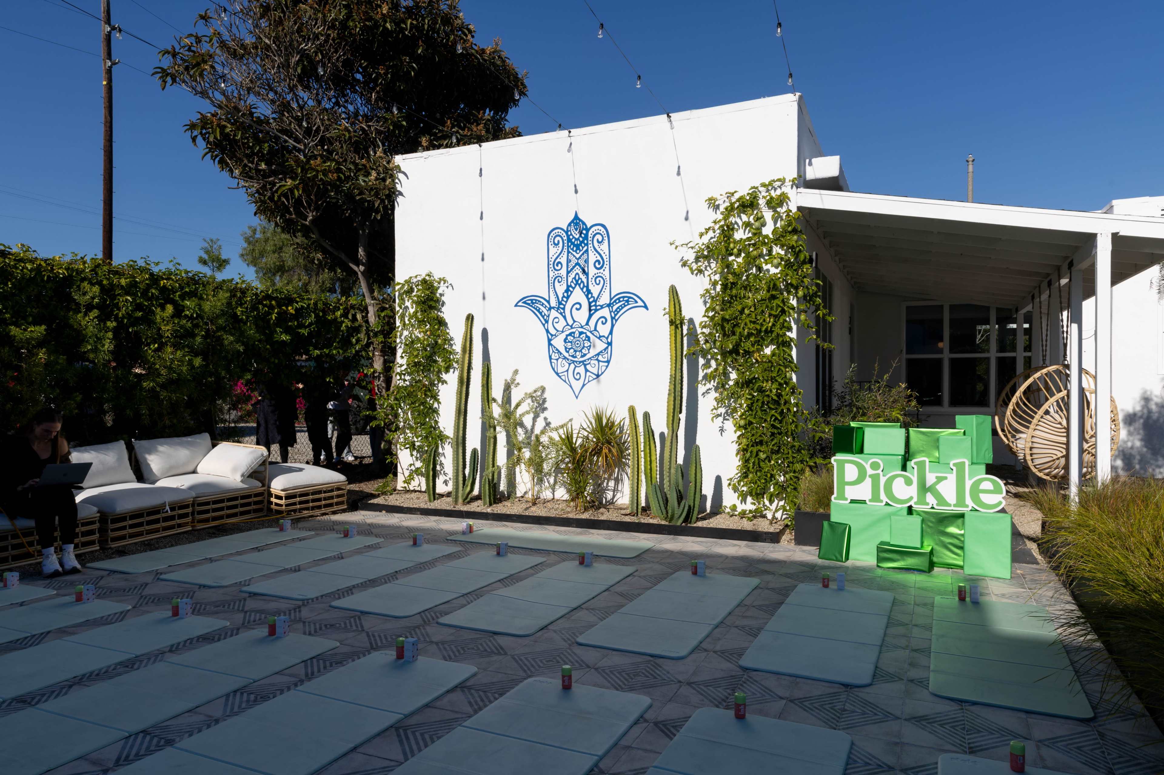 The image shows an outdoor event space featuring a white wall with a blue decorative symbol, surrounded by plants and modern seating, with green signage that reads "Pickle" prominently displayed.
