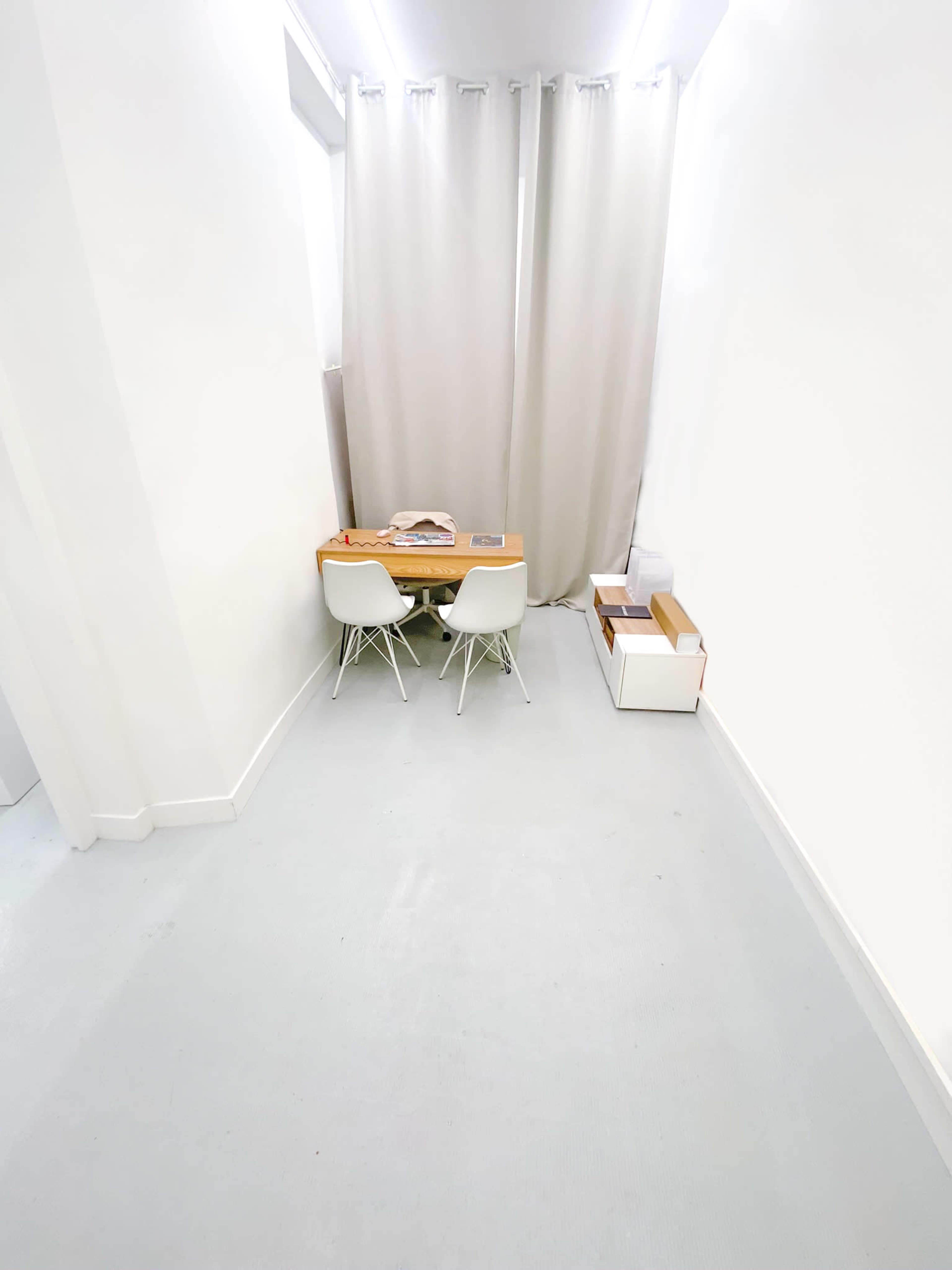 A small, minimalist workspace features a wooden table with two white chairs, situated in a brightly lit hallway beside a large curtain.