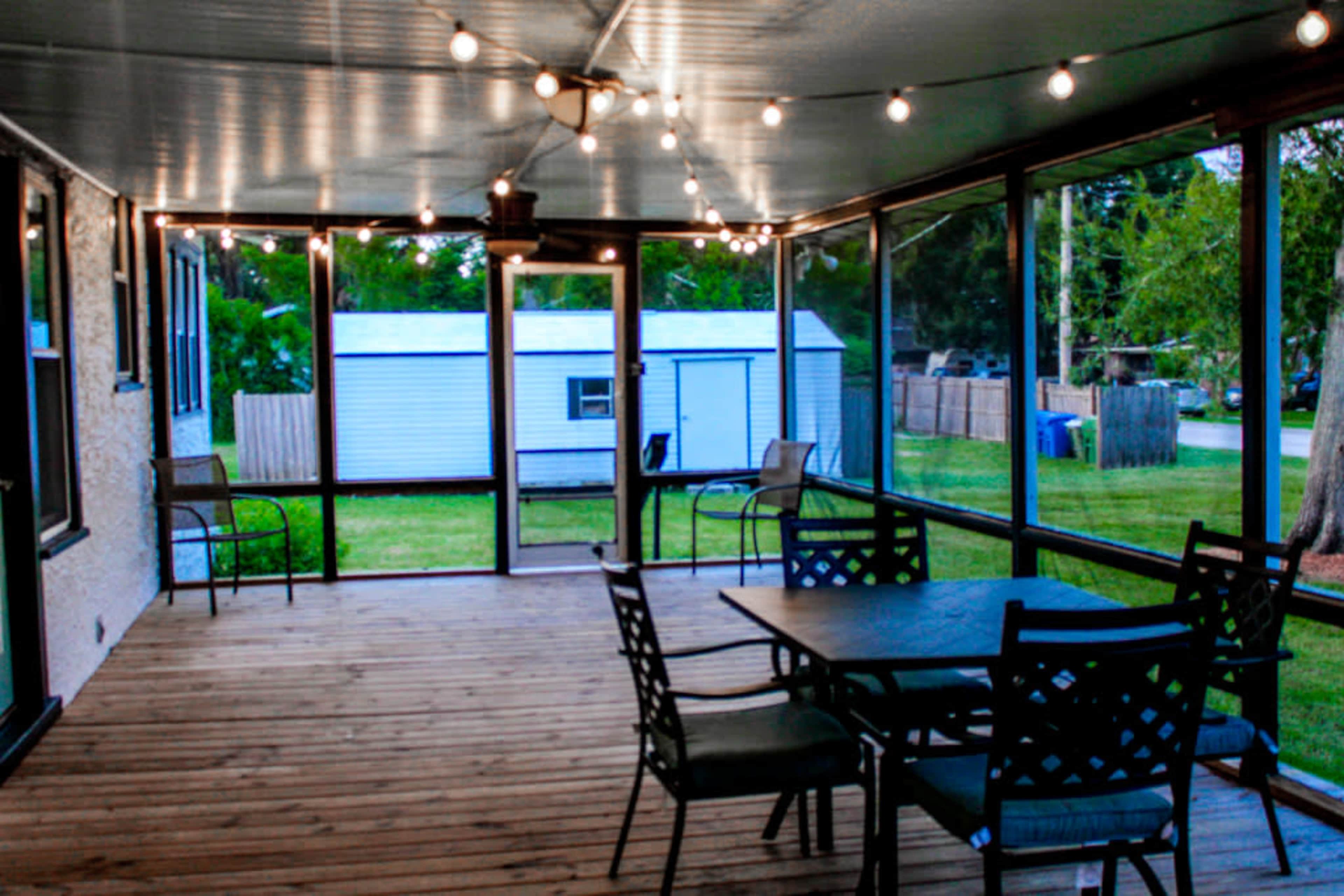 A screened porch features a wooden floor, a dining table with chairs, and string lights overhead, with a view of a grassy yard and a storage shed outside.