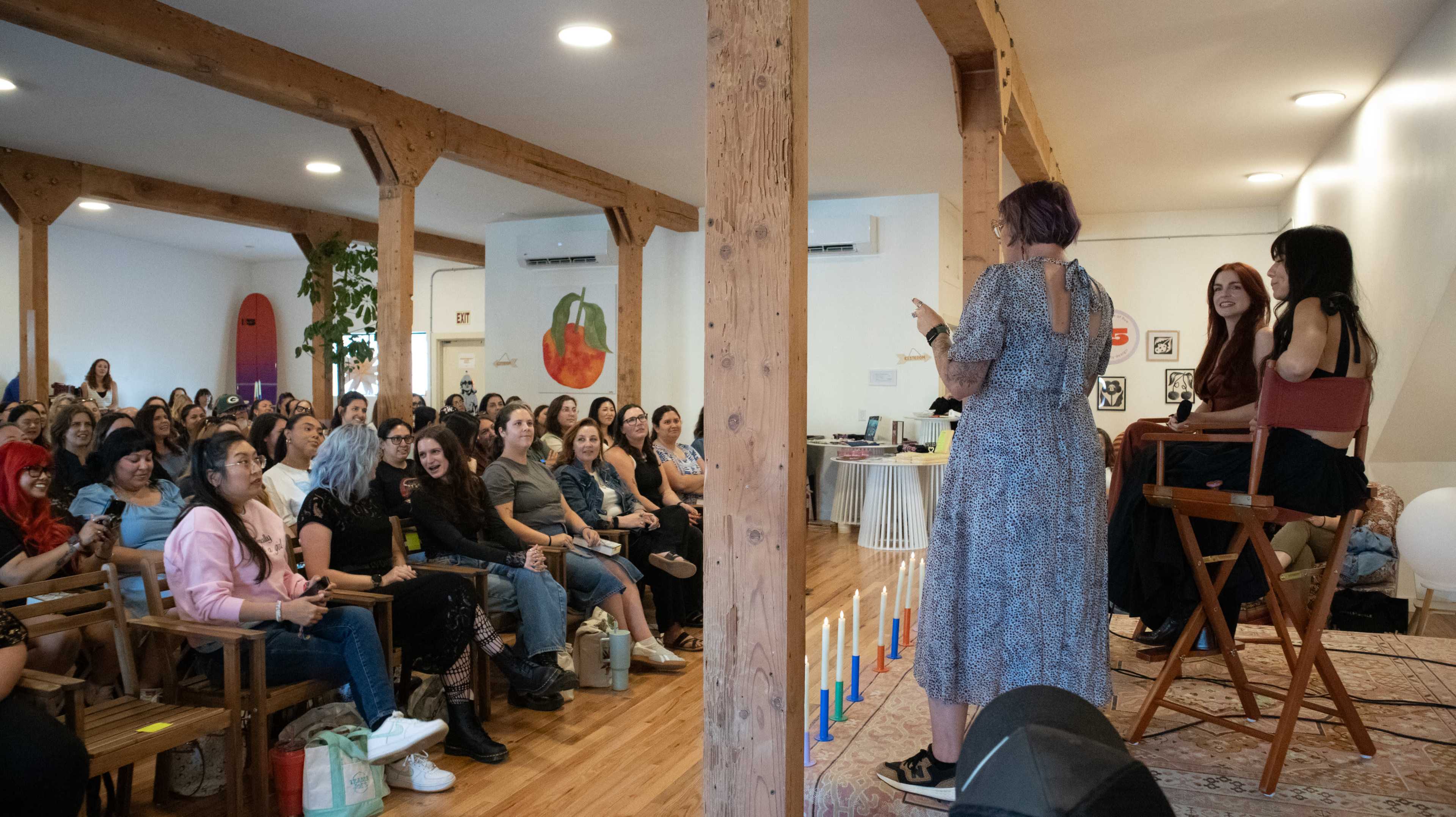A speaker stands on a stage addressing a large audience seated in a well-lit room with wooden beams and a decorative rug.