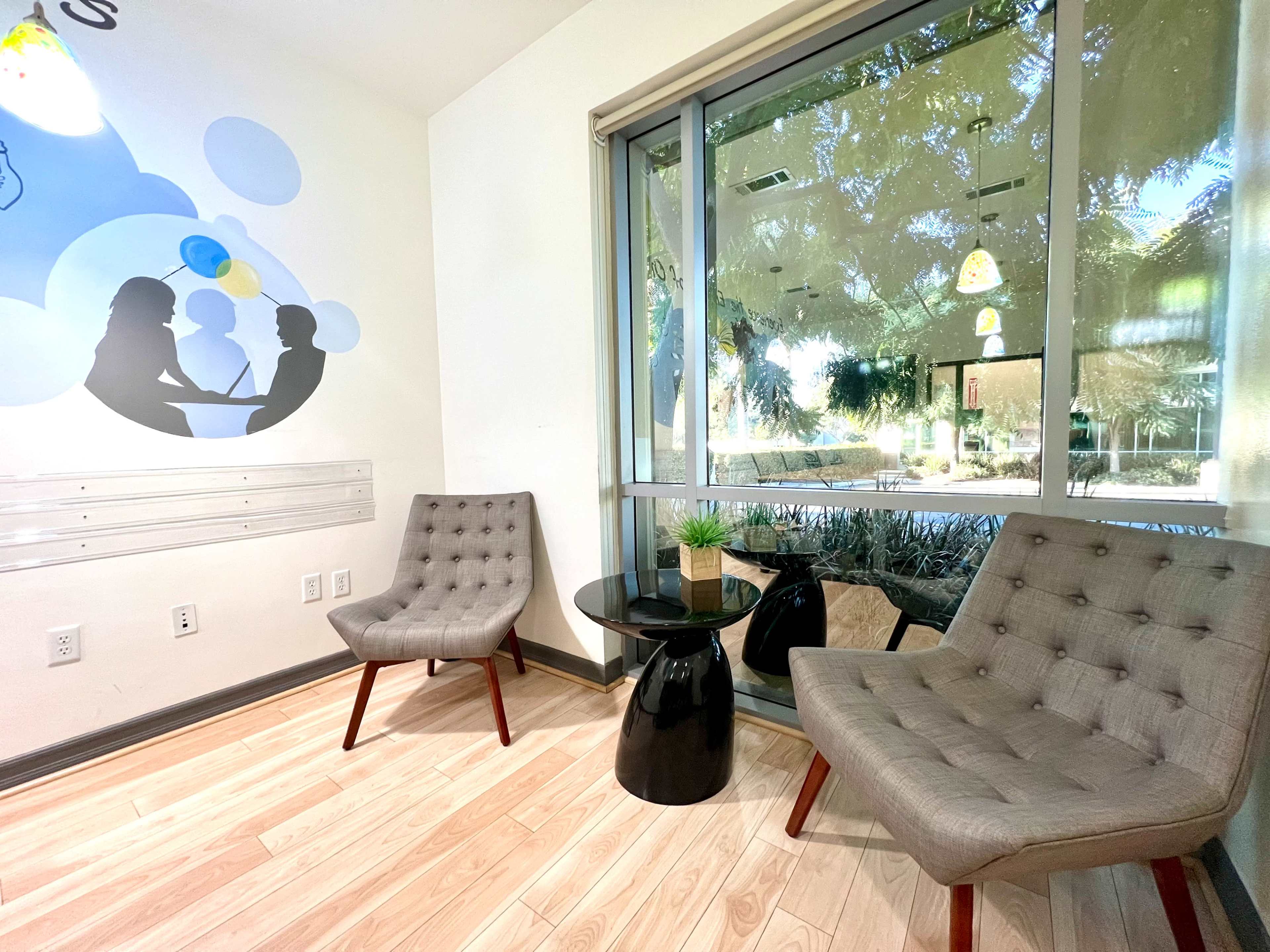 The image shows a seating area with two gray chairs, a black round table, and large windows overlooking an outdoor space.