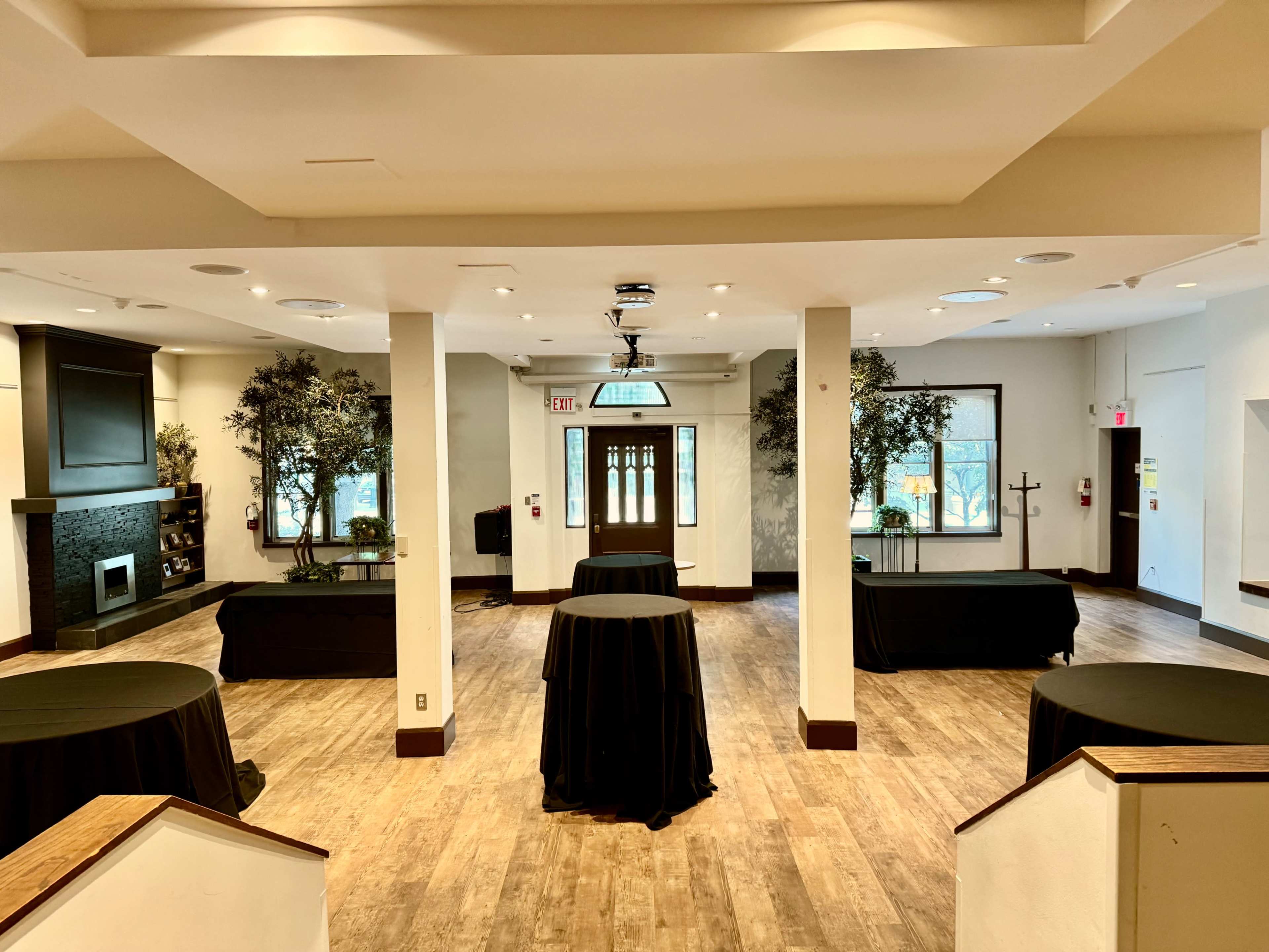 The image shows a spacious, unfurnished event hall with several round tables covered in black cloth, surrounded by trees and a fireplace.