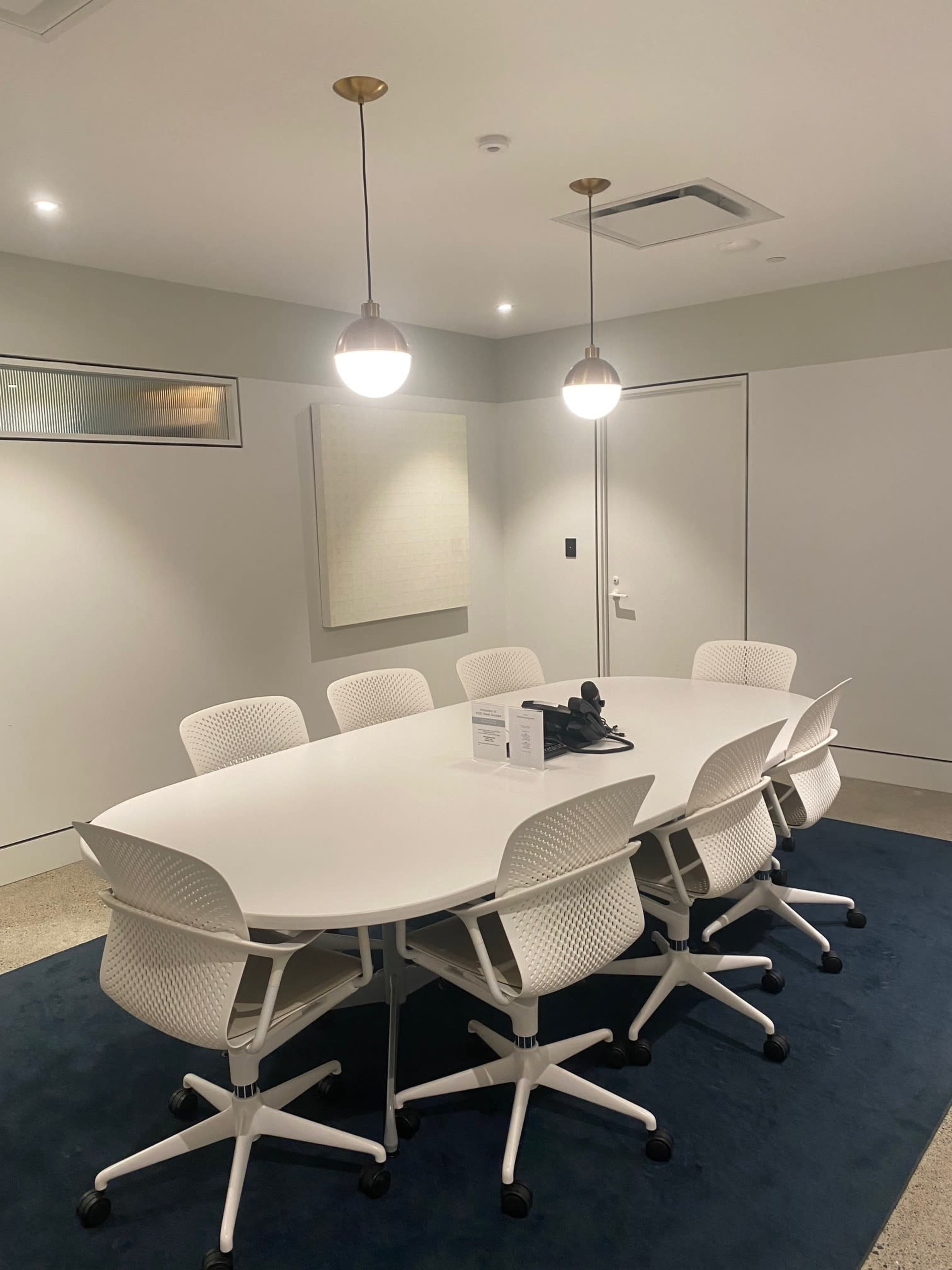 A modern conference room features a white oval table surrounded by eight white rolling chairs, with two pendant lights hanging above and a textured wall panel nearby.