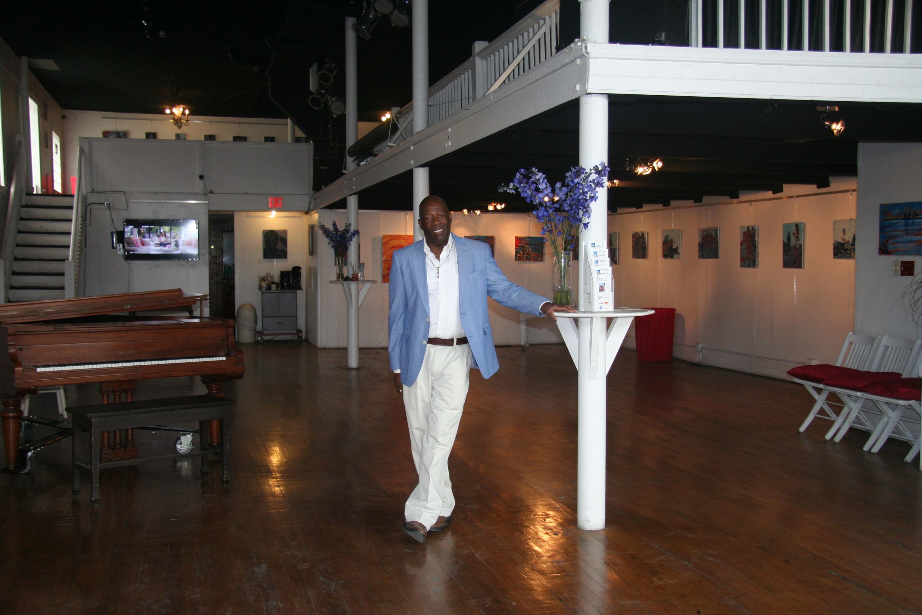A man in a light blue blazer and white pants stands beside a column in an art gallery with wooden floors and a grand piano.
