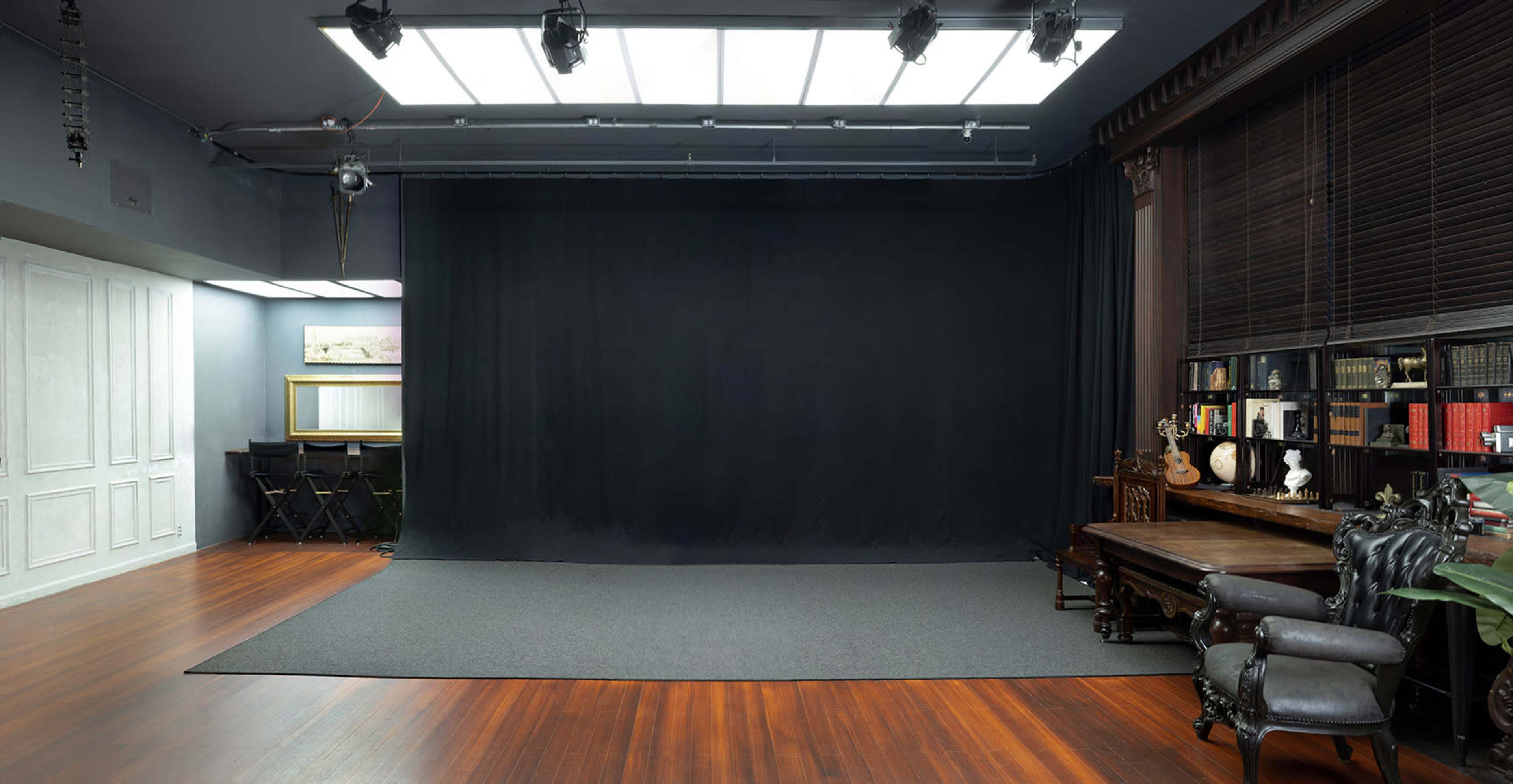 A spacious studio interior featuring a black backdrop, wooden flooring, and a well-furnished area with bookshelves, a table, and chairs.