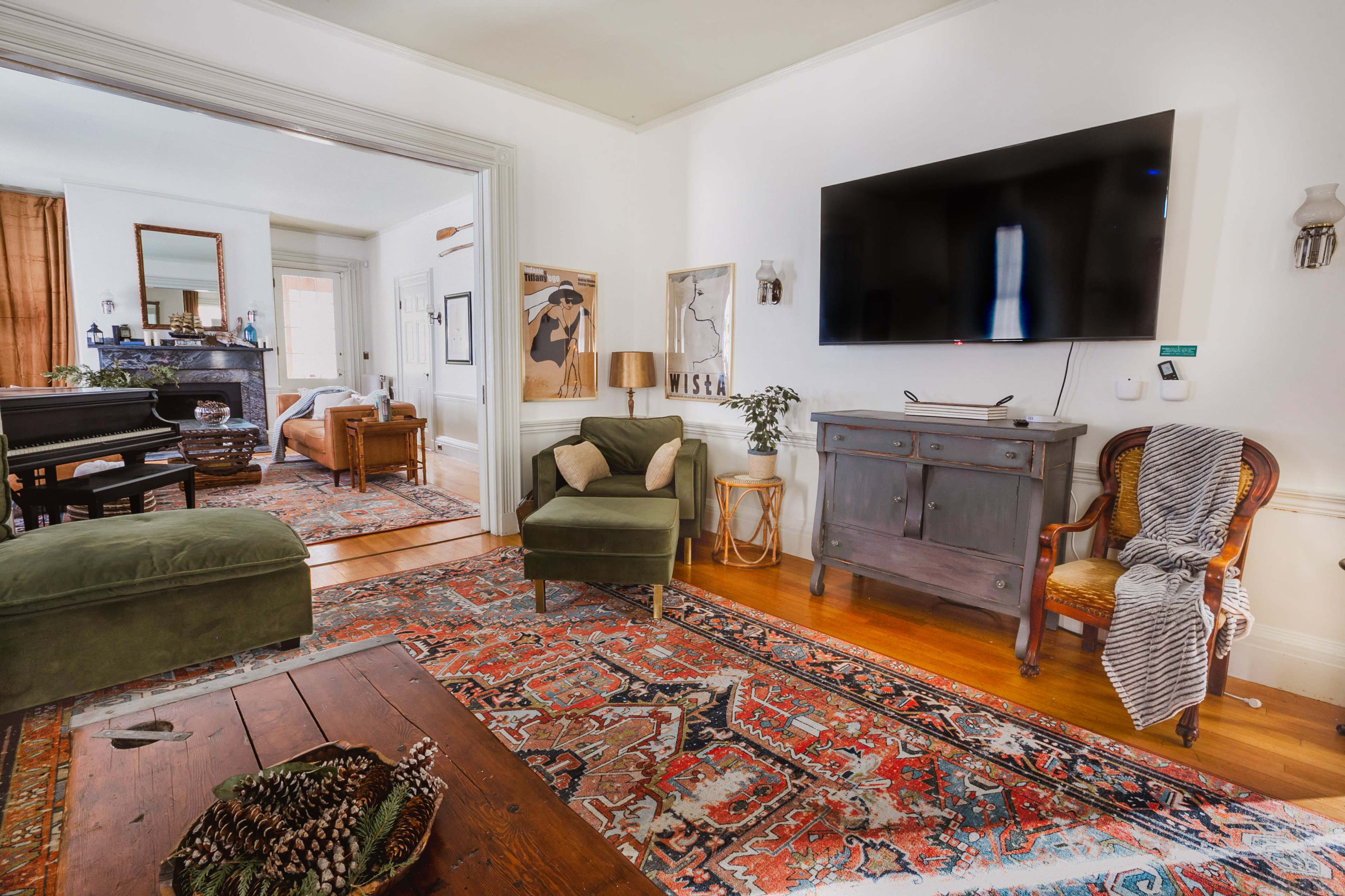 The image shows a cozy living room with a green upholstered chair, a wooden coffee table, and a television mounted on the wall, featuring a large area rug and various pieces of furniture in a bright, airy space.