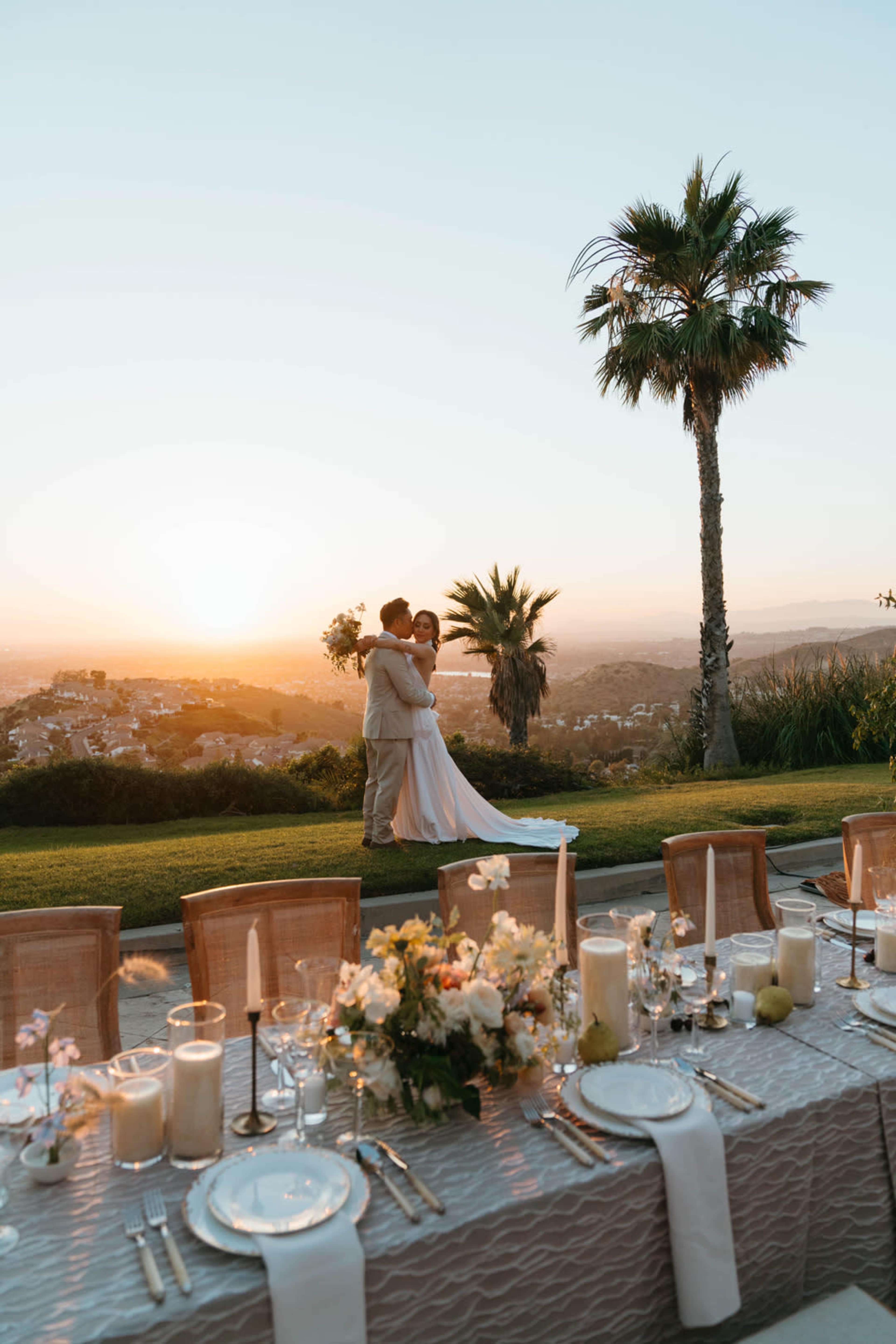 A couple embraces in a wedding setting with a decorated table and a sunset view in the background.