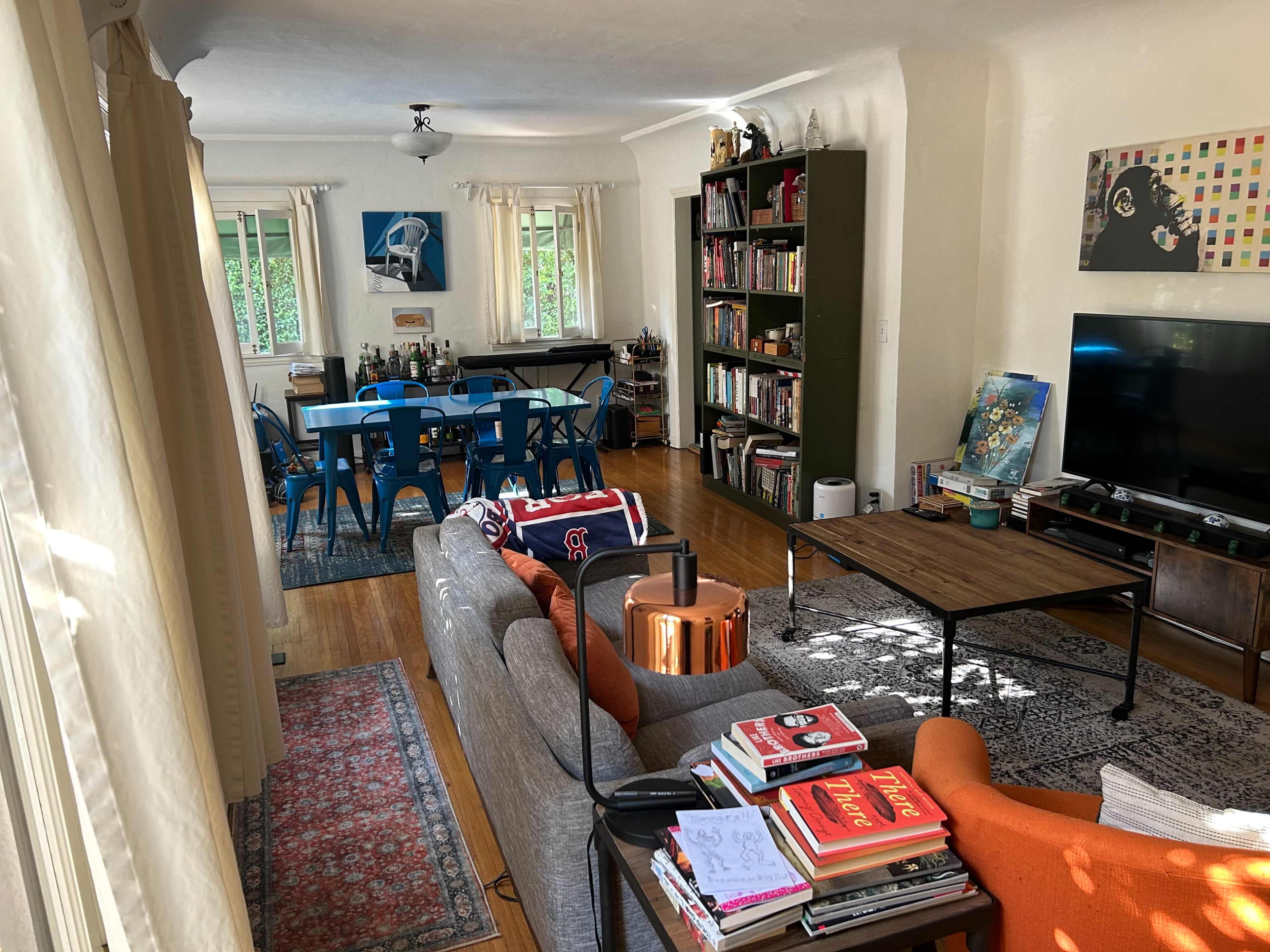 The image shows a living room connected to a dining area, featuring a couch, coffee table, shelves of books, a television, and a dining table with chairs.