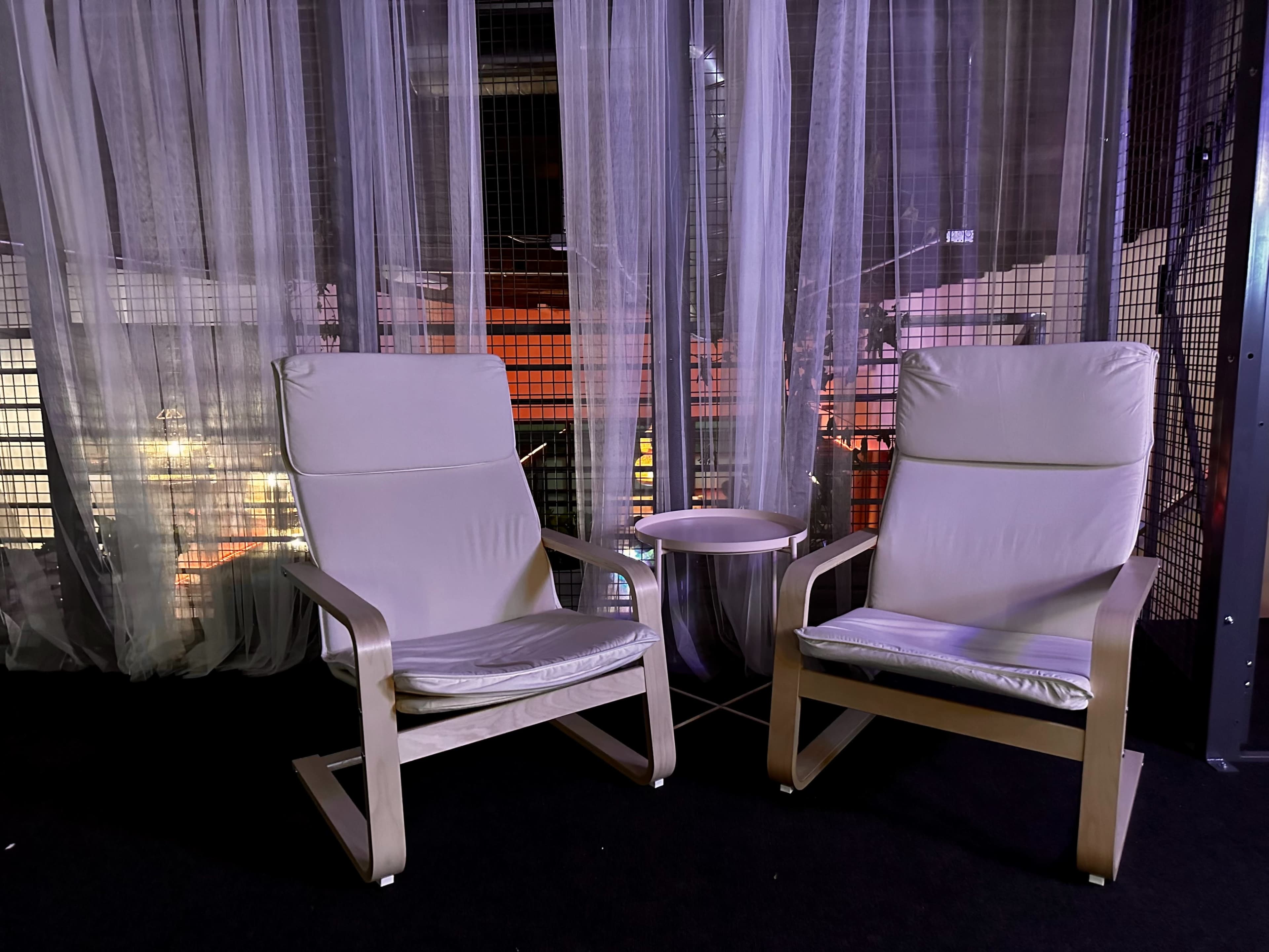 Two modern lounge chairs are positioned beside a small table, set against a backdrop of sheer curtains in a dimly lit environment.