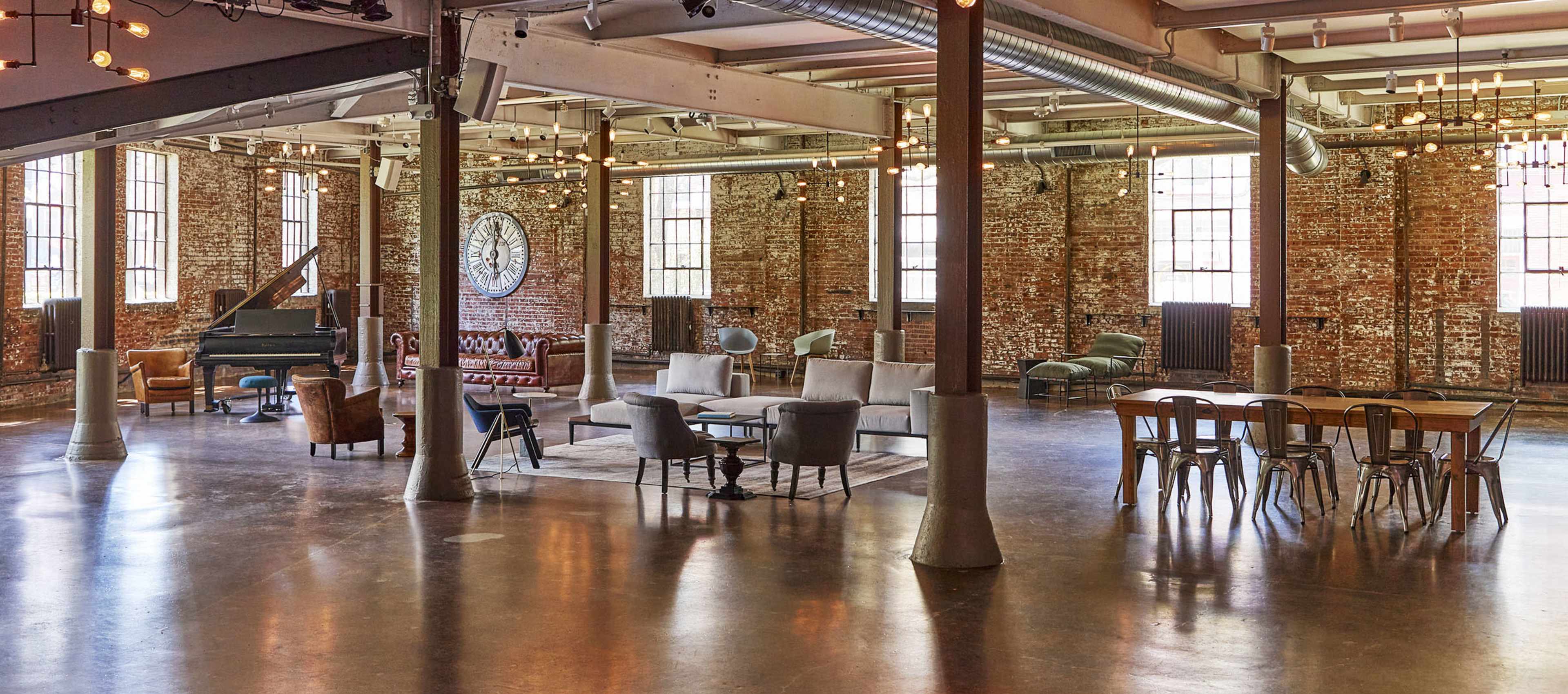The image shows a spacious industrial loft with exposed brick walls, large windows, elegant furniture, and various seating arrangements.