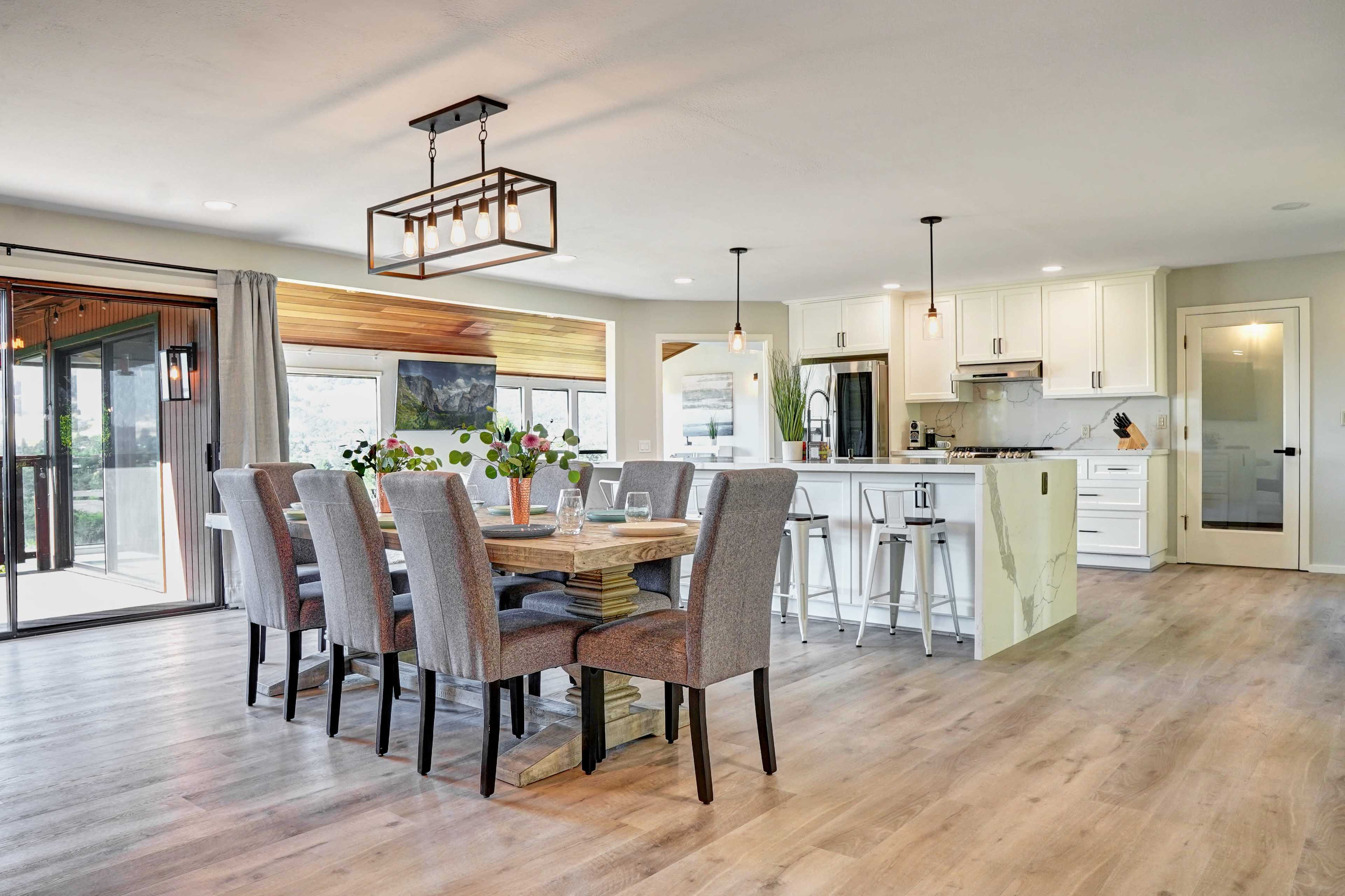 The image shows a modern dining area with a wooden table surrounded by gray upholstered chairs, adjacent to a bright kitchen featuring white cabinets and bar stools.