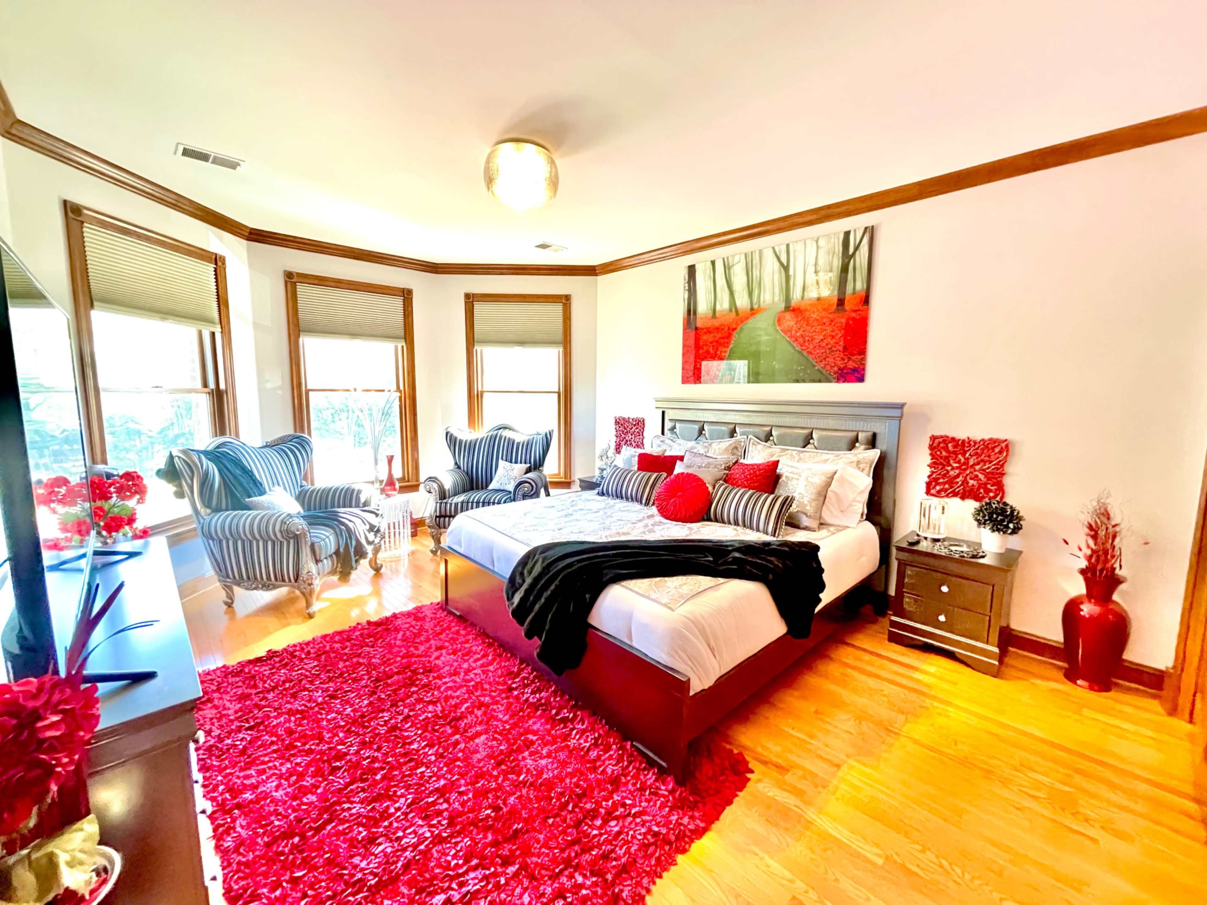 The bedroom features a large bed with red and white bedding, two striped armchairs, a red area rug, and artwork depicting a forest scene on the wall.