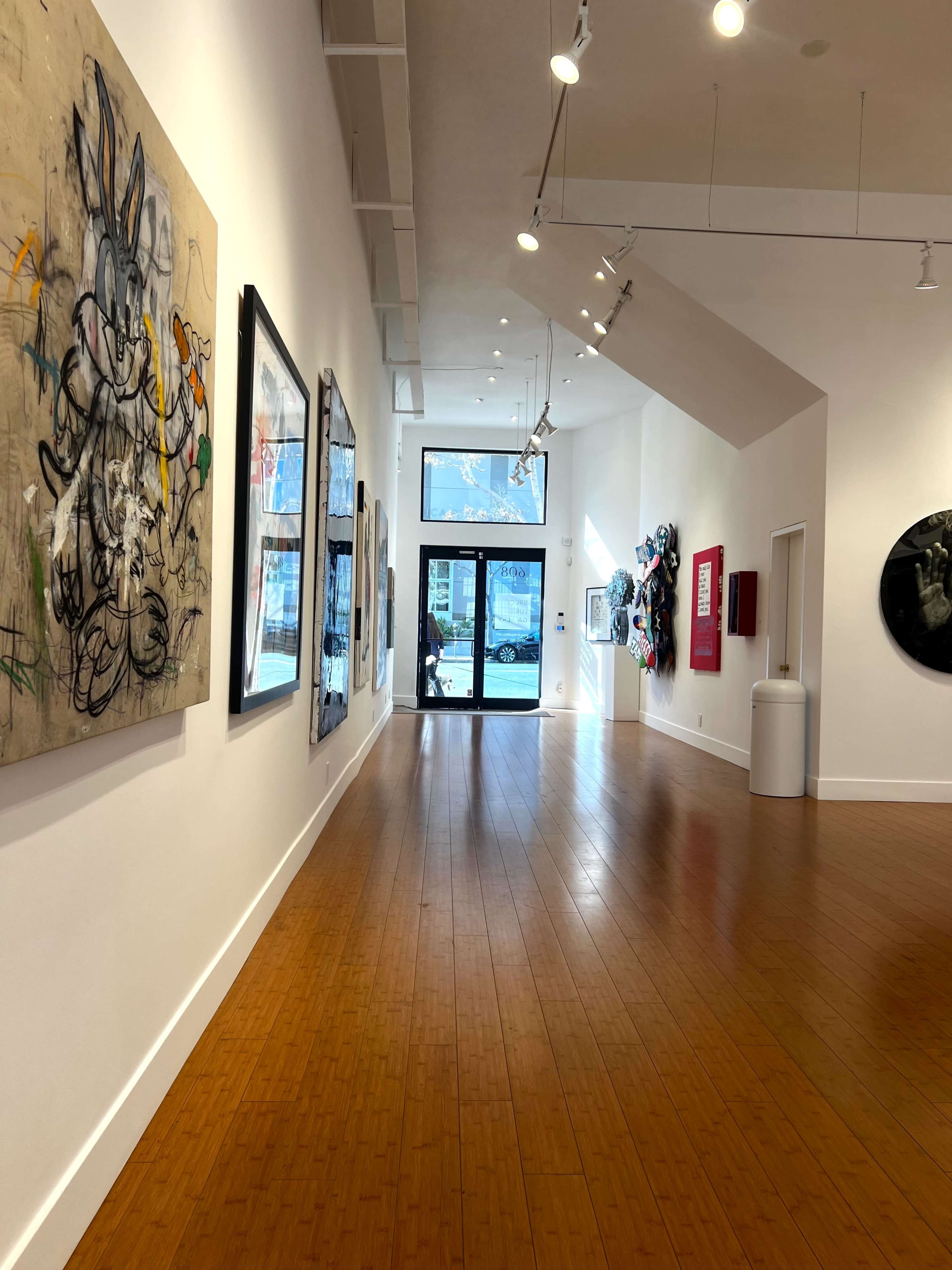 The image shows a spacious art gallery with several framed artworks displayed along the white walls and a large glass entrance at the far end.