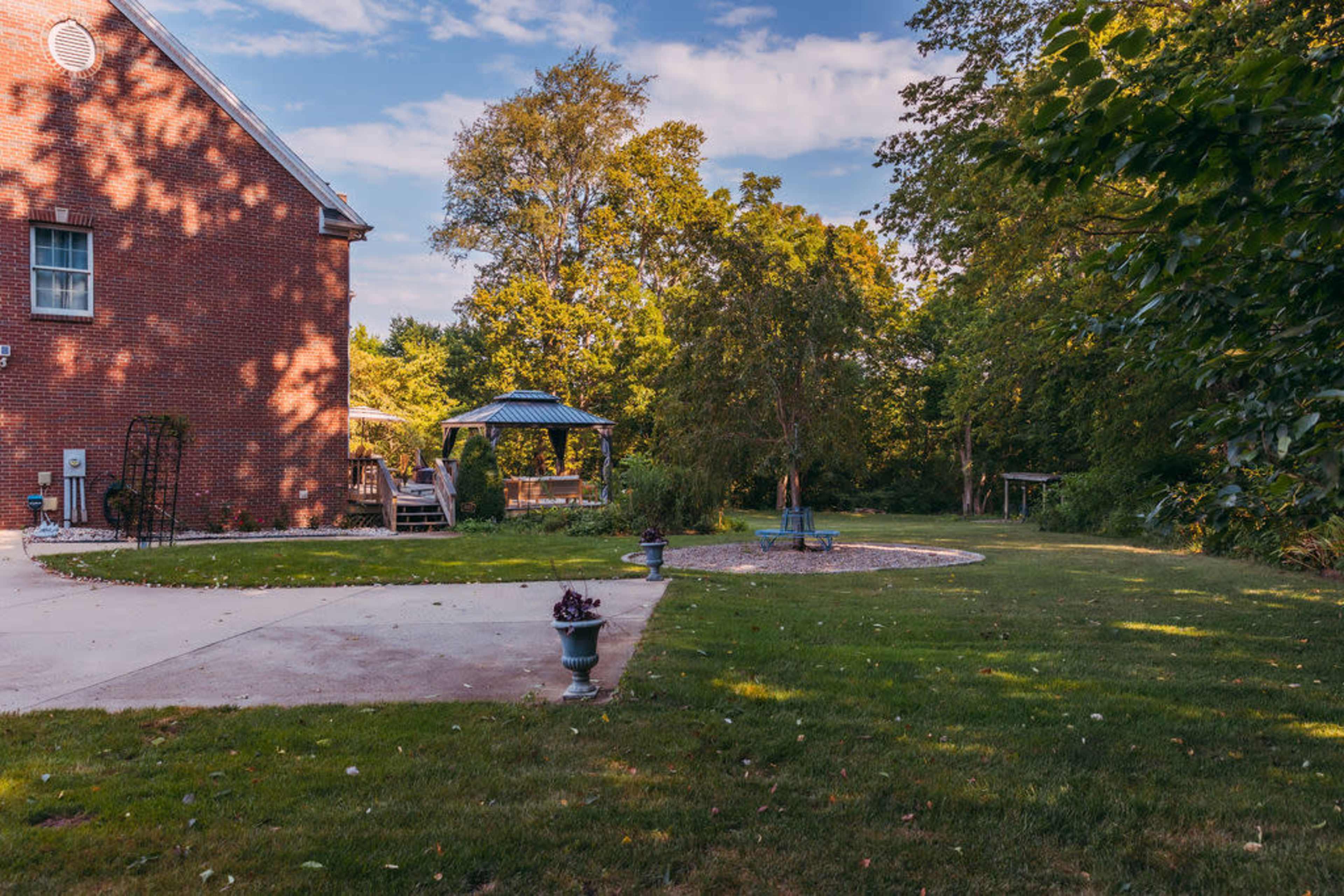 A brick house with a patio, surrounded by a manicured lawn and several trees in a serene outdoor setting.