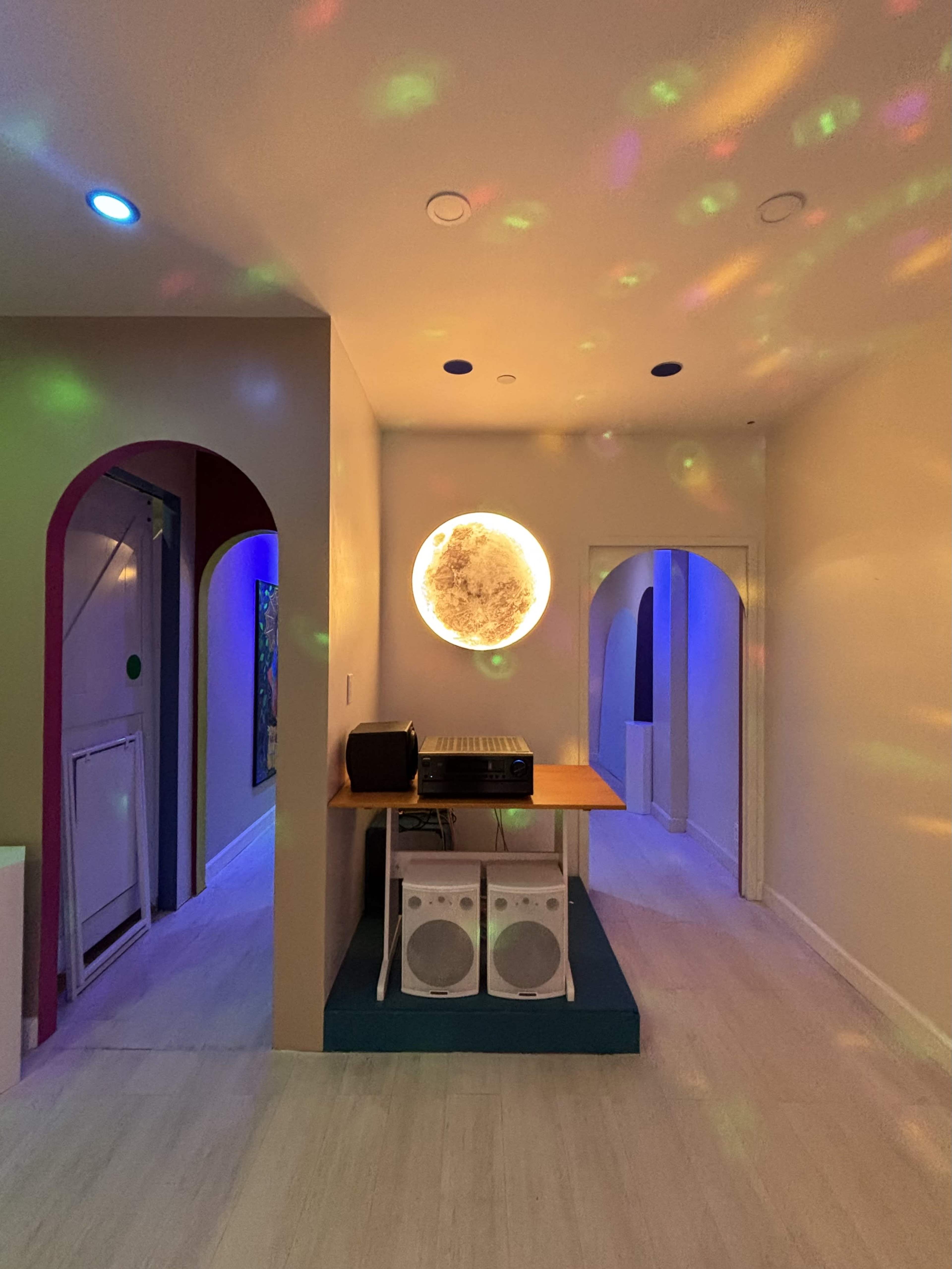 A vibrant room features a DJ setup with speakers and colorful light reflections on the walls, illuminated by a large circular light fixture.