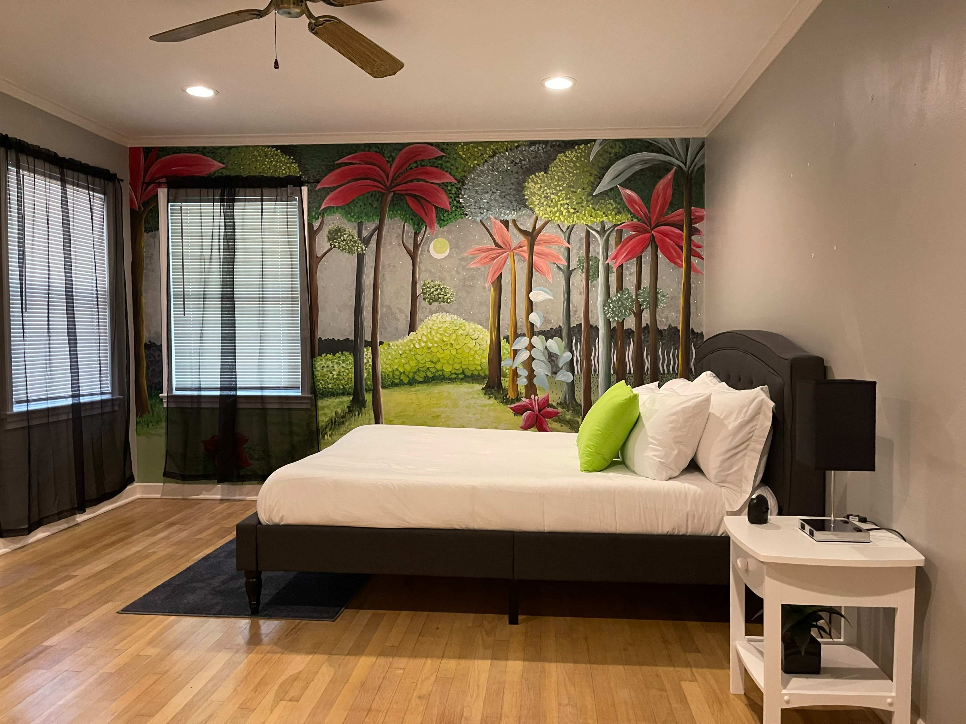 A bedroom features a large bed with white bedding against a mural of a tropical landscape and large windows with sheer black curtains.
