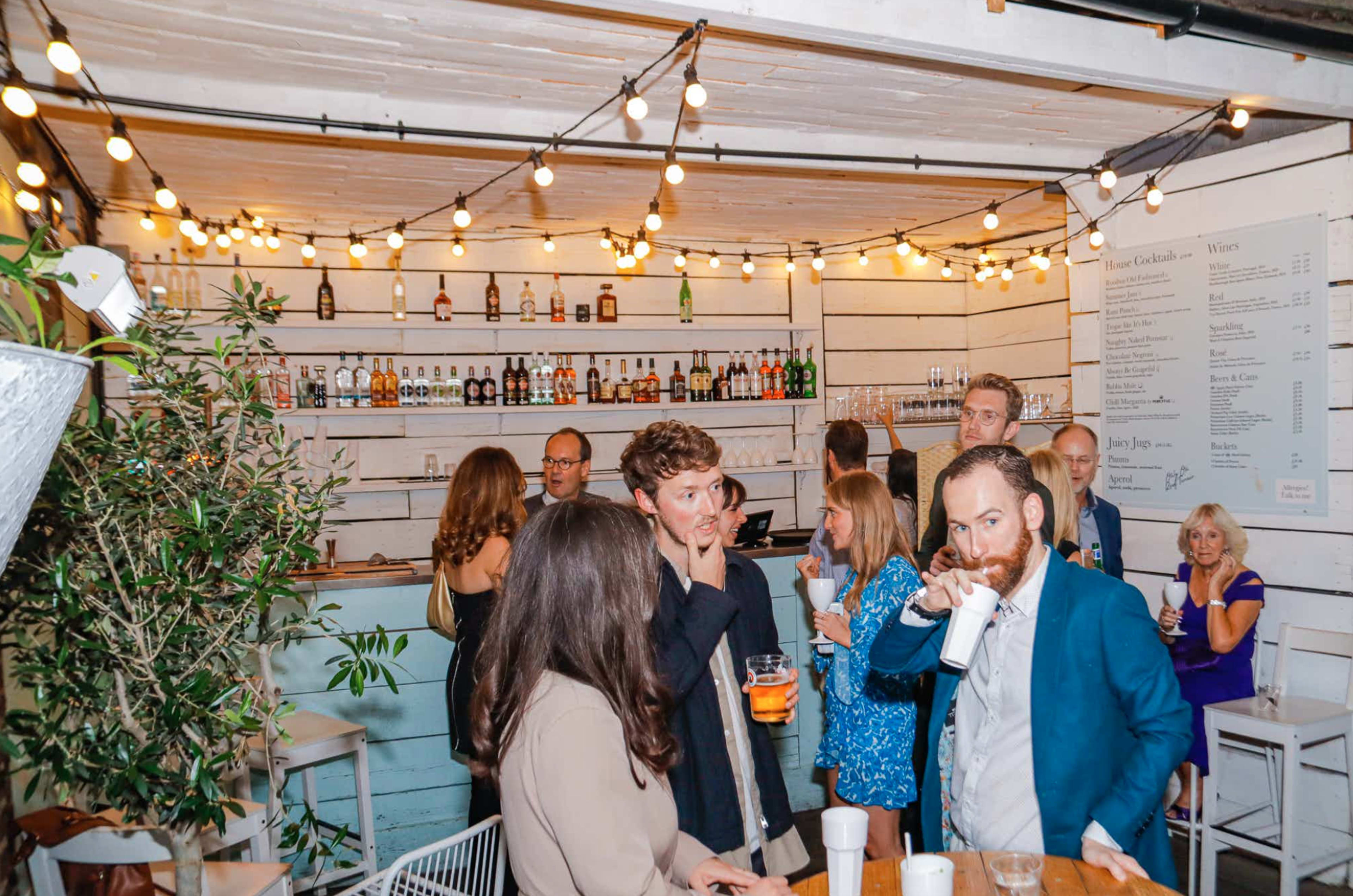 A group of people socialize in a bar with string lights hanging above and shelves lined with various bottles in the background.