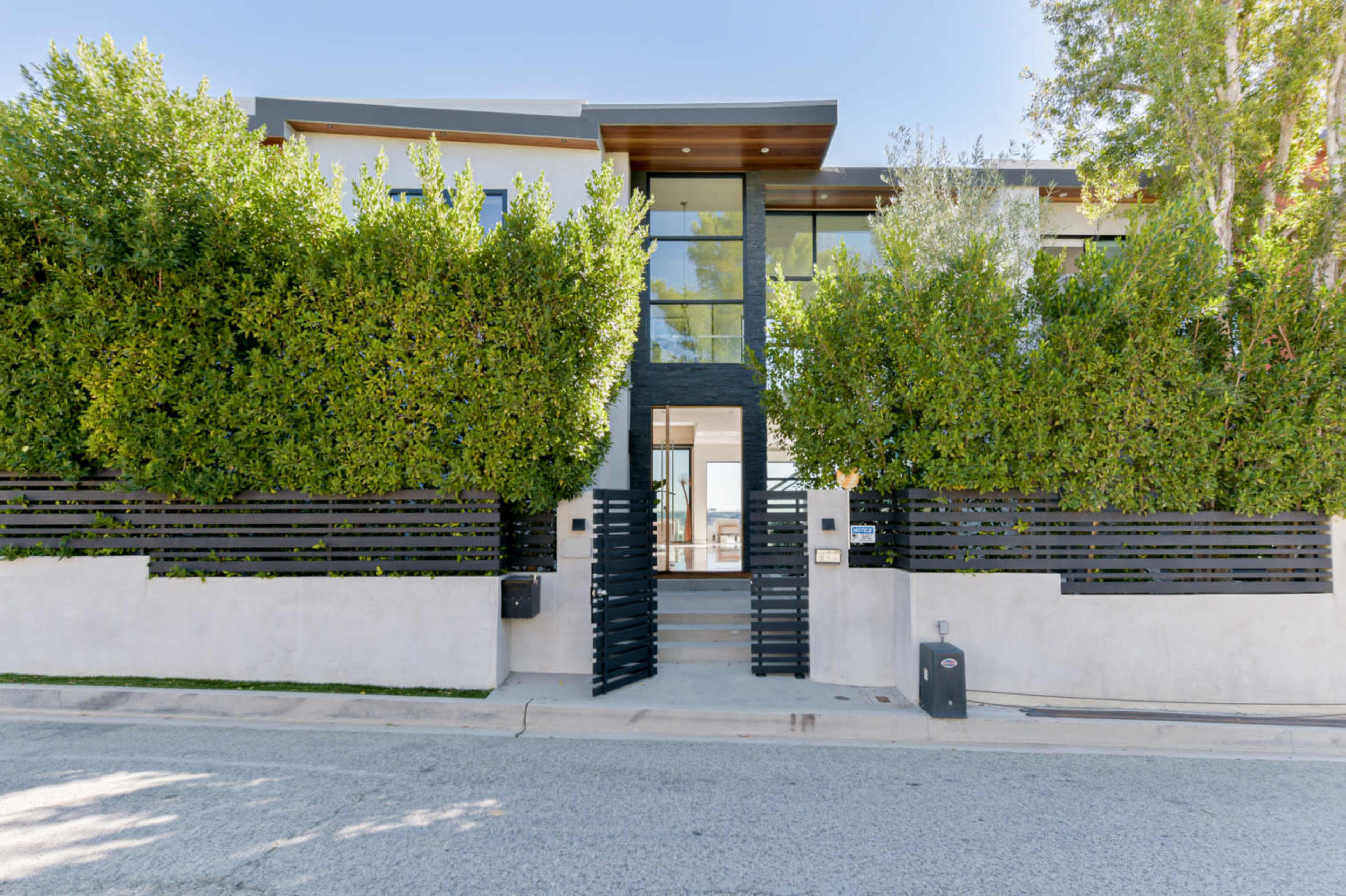 A modern home with large glass windows is surrounded by tall green hedges and features a minimalist black and white facade.