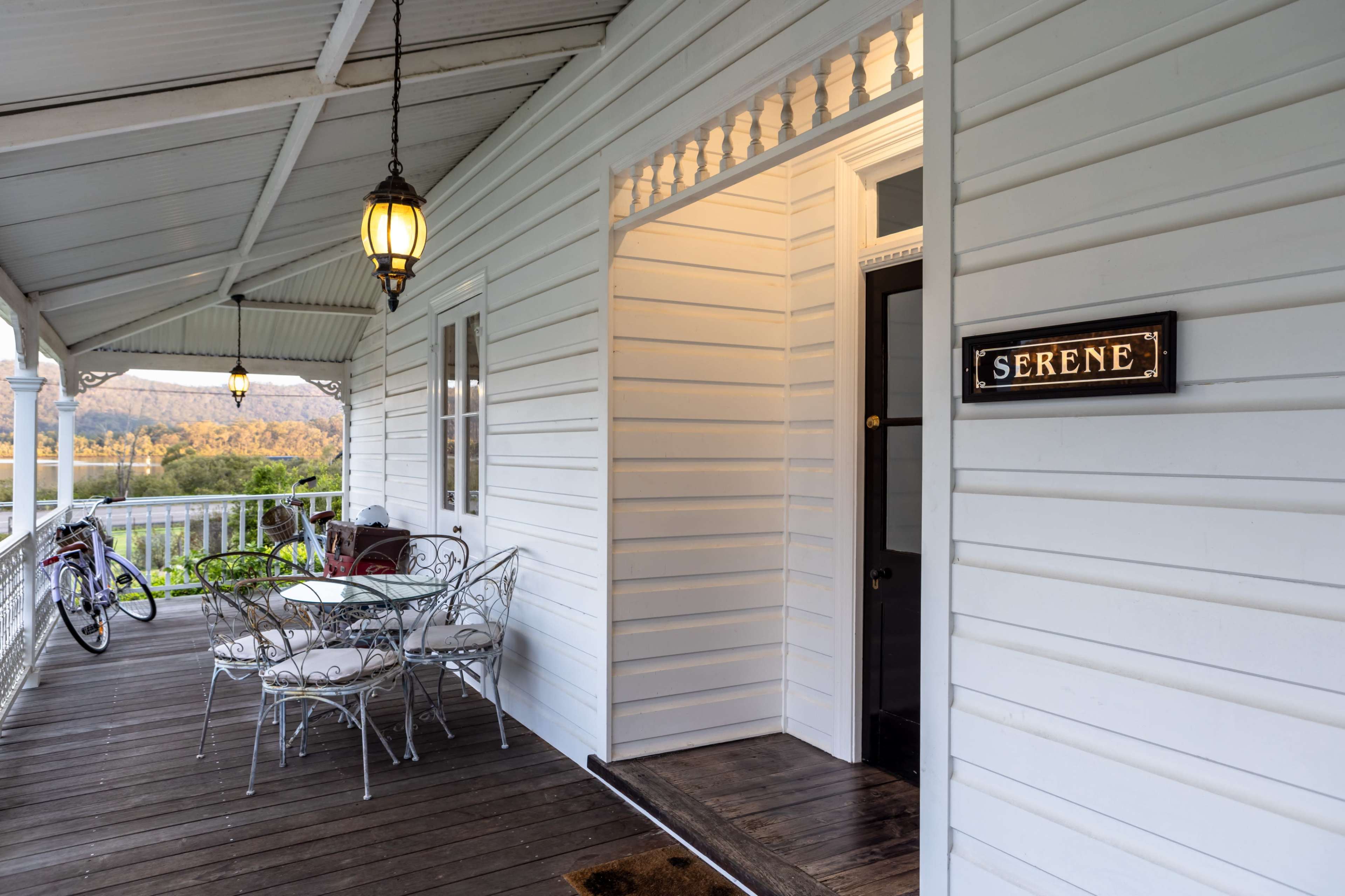 The image shows a veranda with a metal table and chairs, a bicycle hanging on the wall, and a sign labeled "SERENE" next to a doorway.