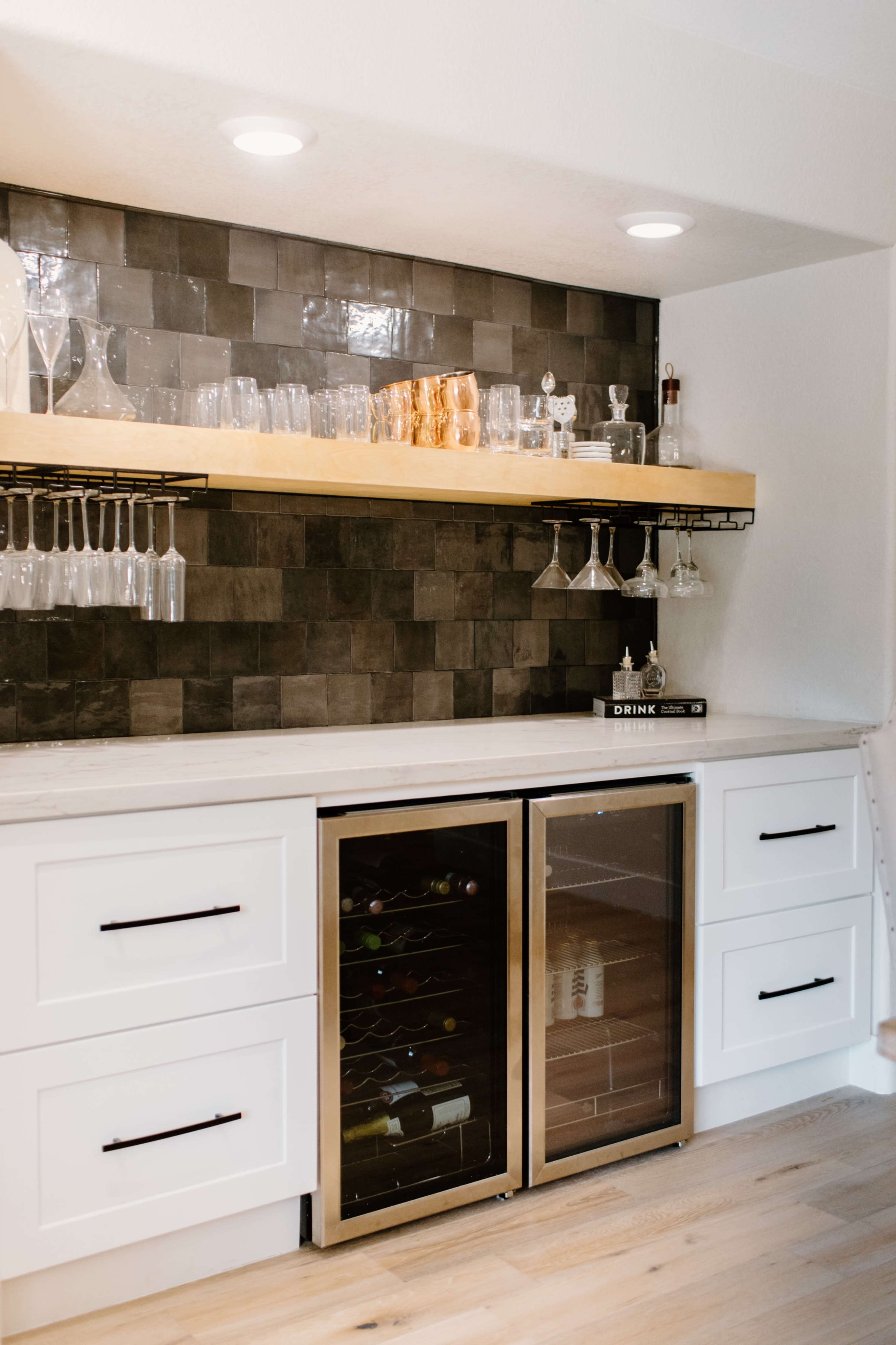 A modern kitchen bar area with a black tiled backsplash, a wooden shelf holding glassware, and two wine refrigerators below a countertop.