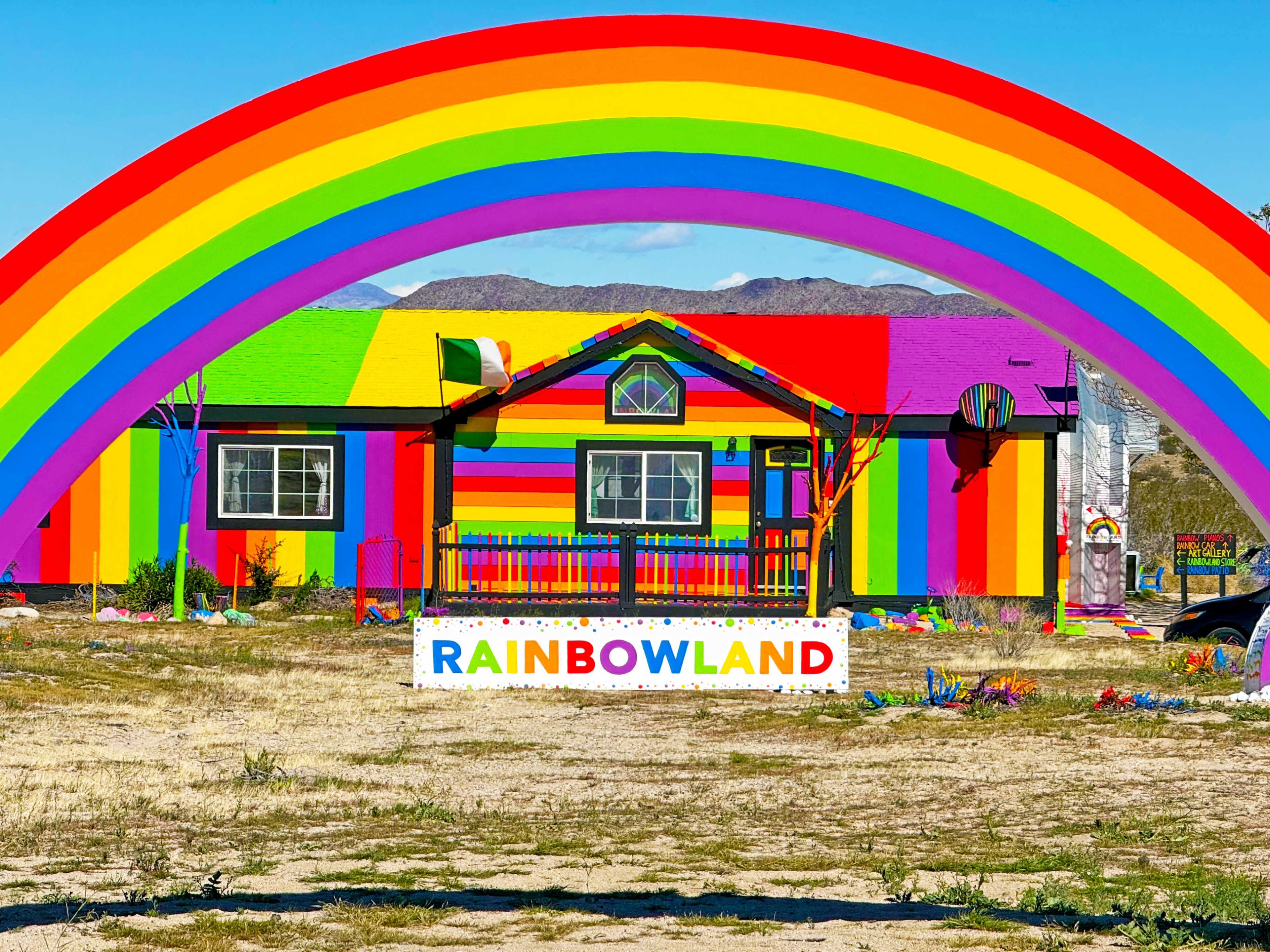 A brightly painted house with rainbow stripes stands under a large rainbow arch, featuring a sign that reads "RAINBOWLAND."