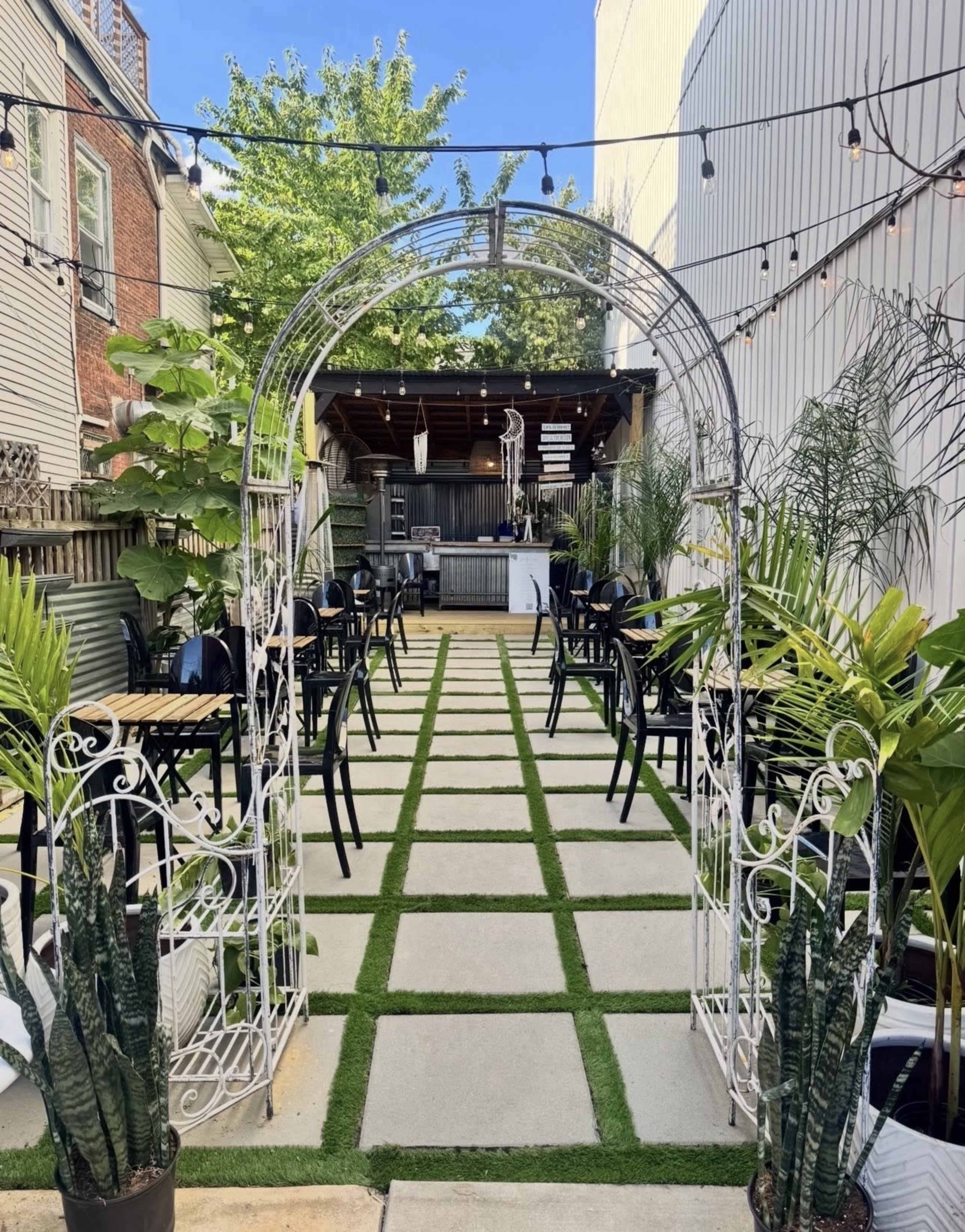 The image shows a landscaped outdoor seating area with a pathway lined with grass and a white archway leading to a bar at the back.