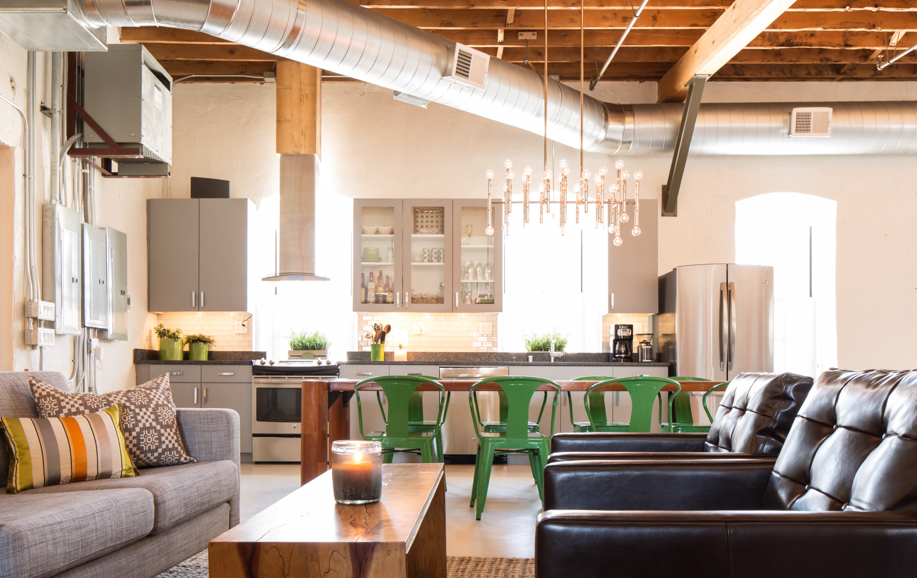 A modern kitchen and living area features stainless steel appliances, green metal chairs, and wooden furniture under exposed beams and ductwork.